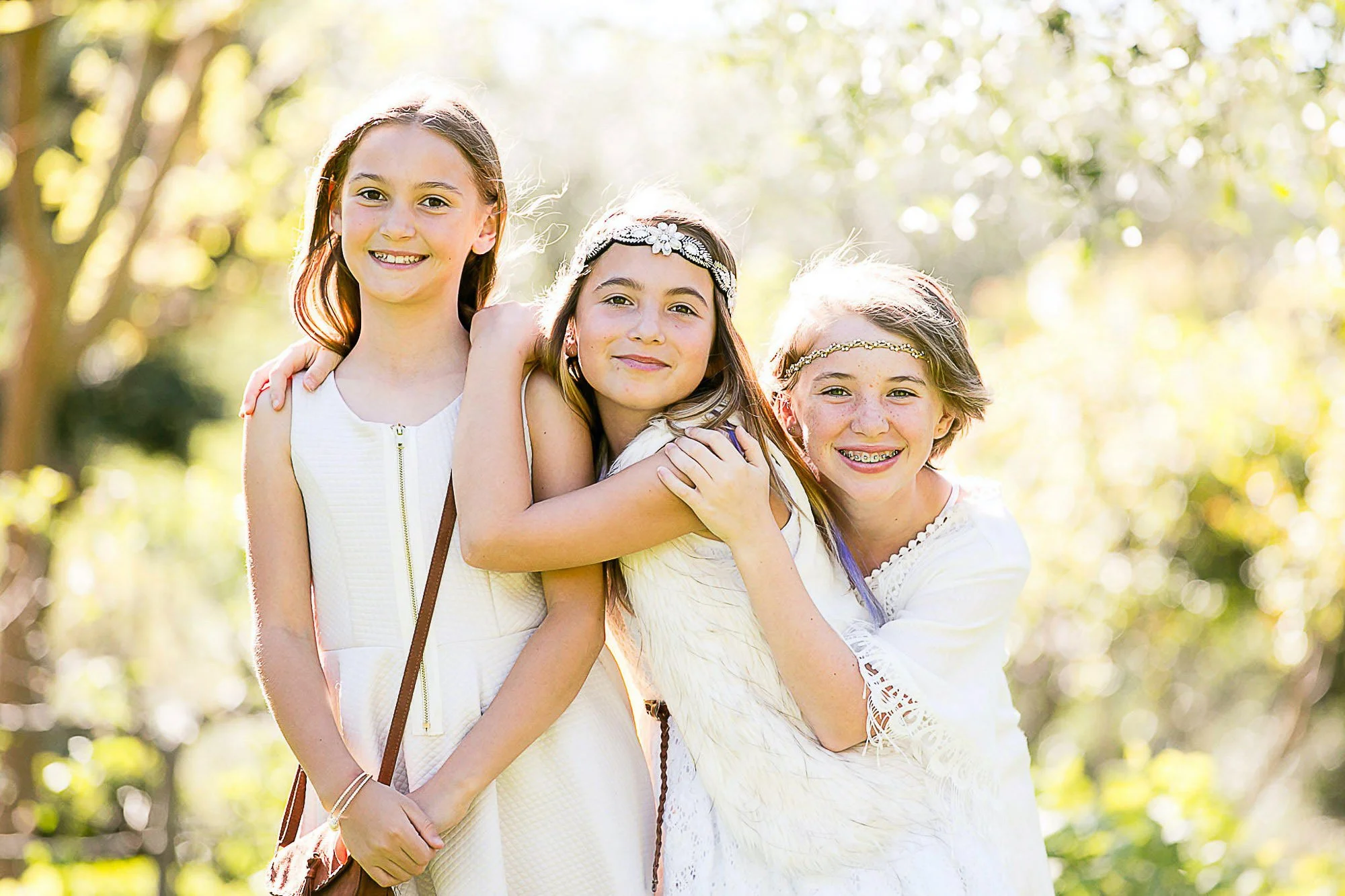 Four young girls smiling in a sunny outdoor setting with trees and bokeh background, wearing white dresses and accessories, embracing each other.