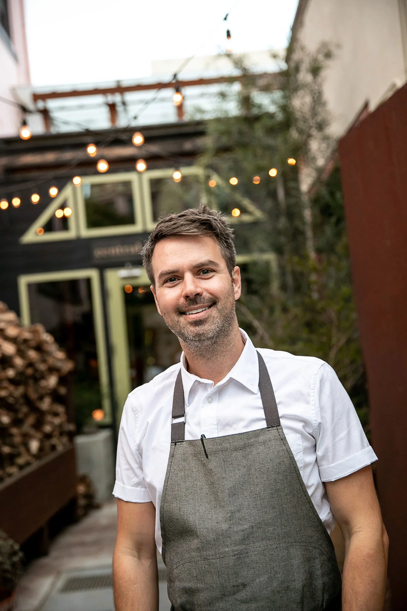 A smiling man with short brown hair, wearing a white shirt and a gray apron, stands outside in front of a modern building with string lights and a large window, during daytime.