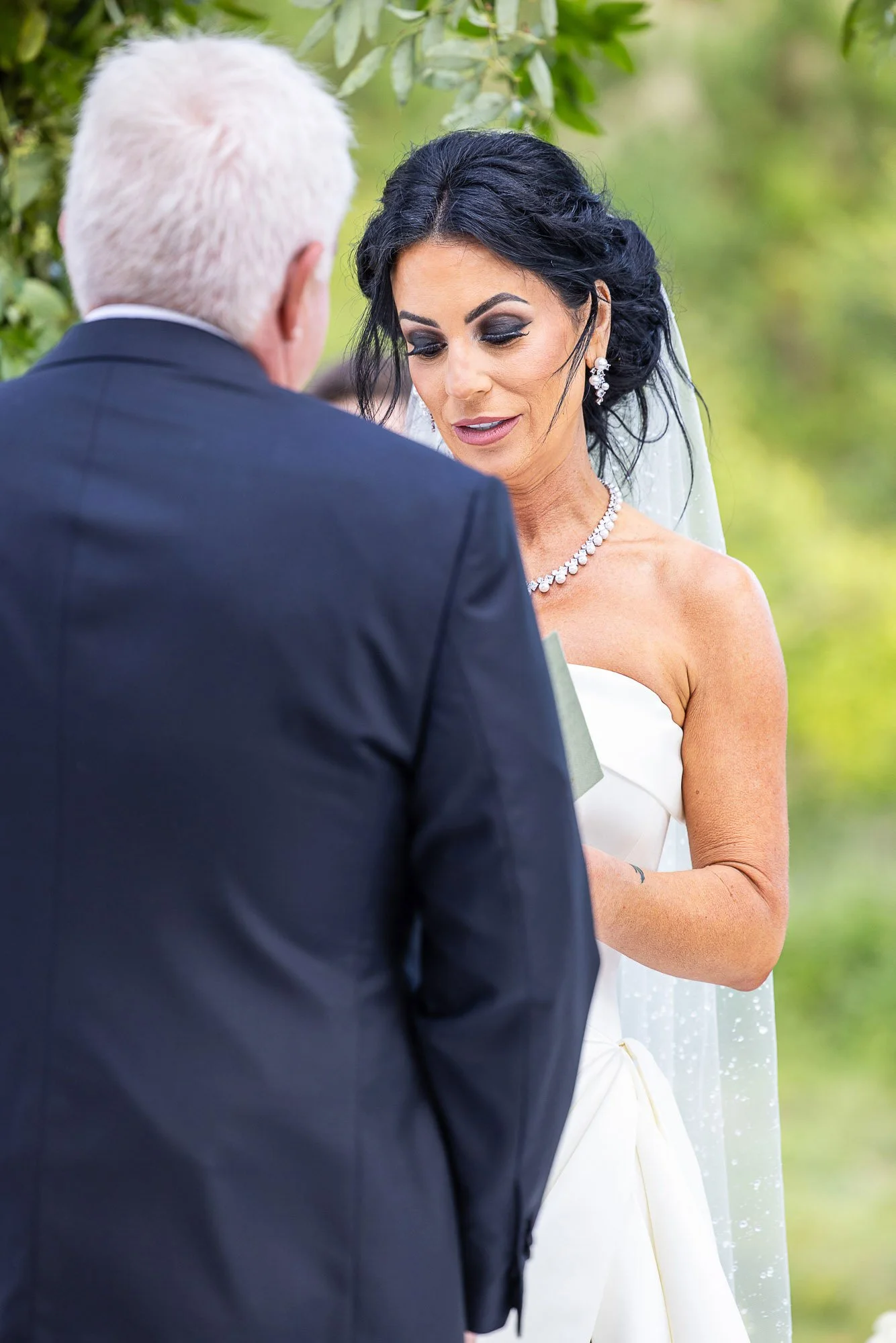 A bride in a white wedding dress with dark hair and makeup, wearing jewelry, standing outside and looking at a man in a dark suit with white hair, likely during a wedding ceremony at the Beach and Tennis Club in Pebble Beach.