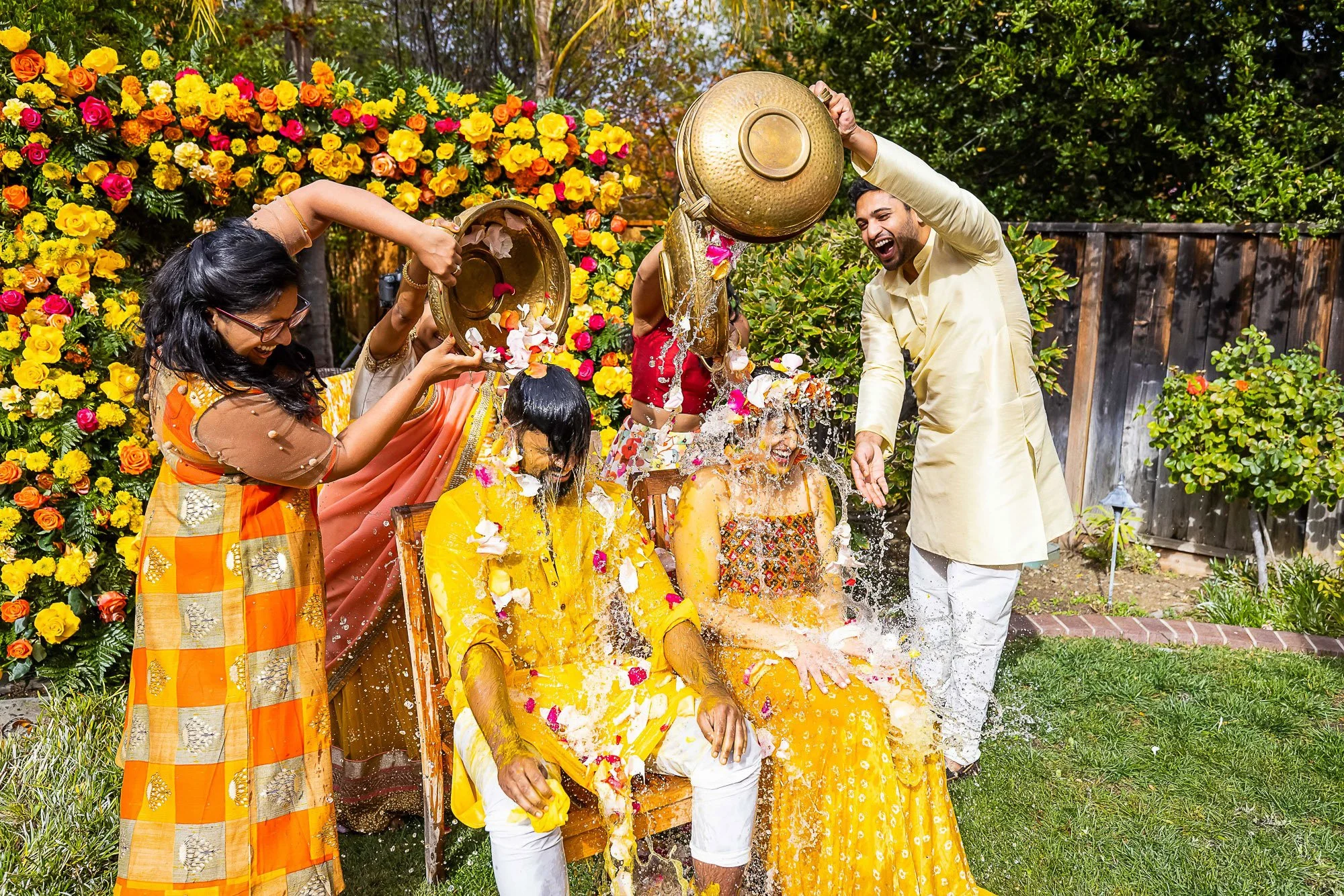 People celebrating an Indian wedding by splashing brightly colored yellow, orange, pink, and white flower petals and water on each other. Two women are sitting on a yellow decorated chair, covered in yellow and orange fabric, while men stand nearby. 
