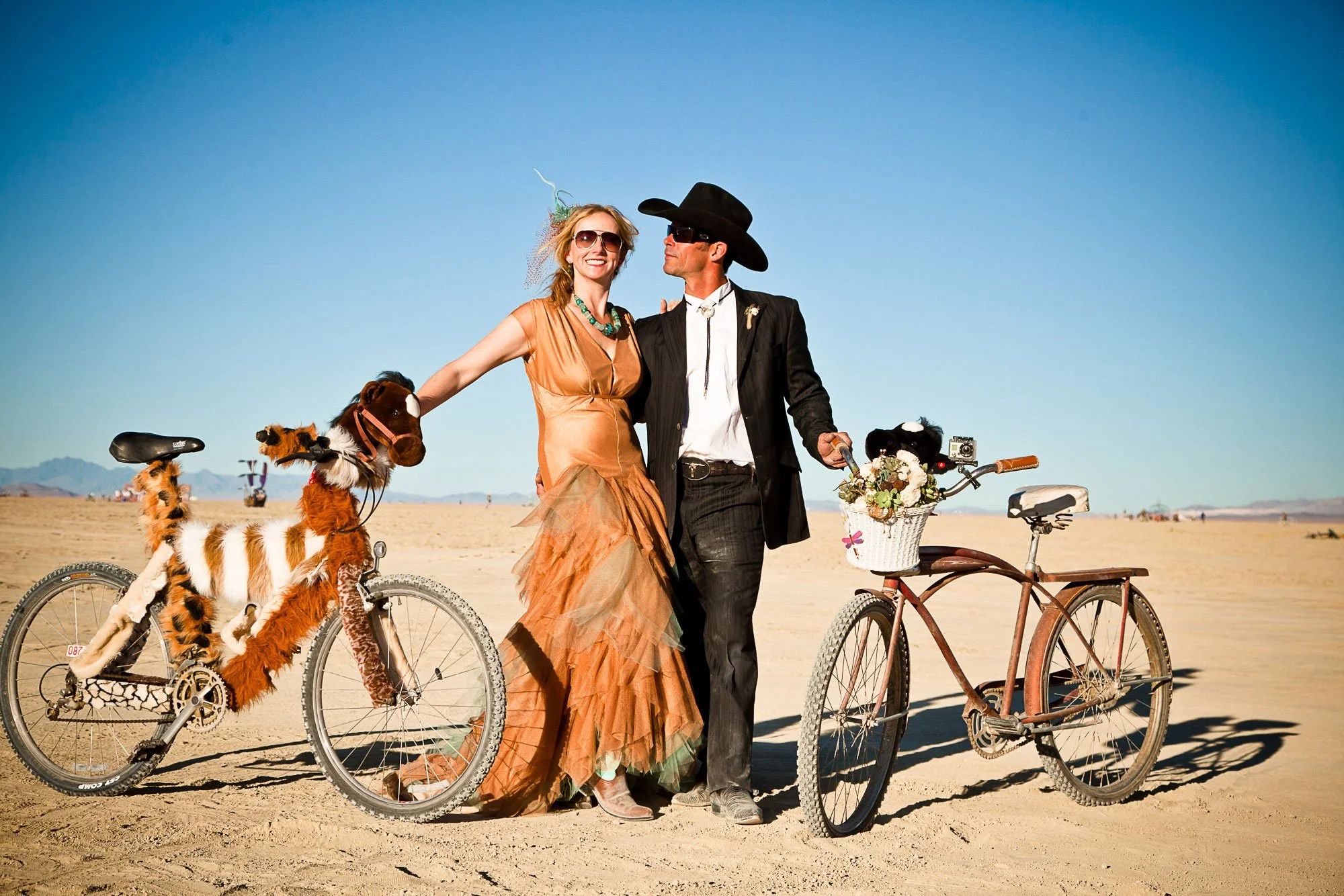 Tara Arrowood and Travis Pynn's wedding at Burning Man. Travis is wearing a wide-brimmed hat stand together outdoors in a desert landscape. Two bicycles, each decorated with plush animal toys and flowers, are in front of them.