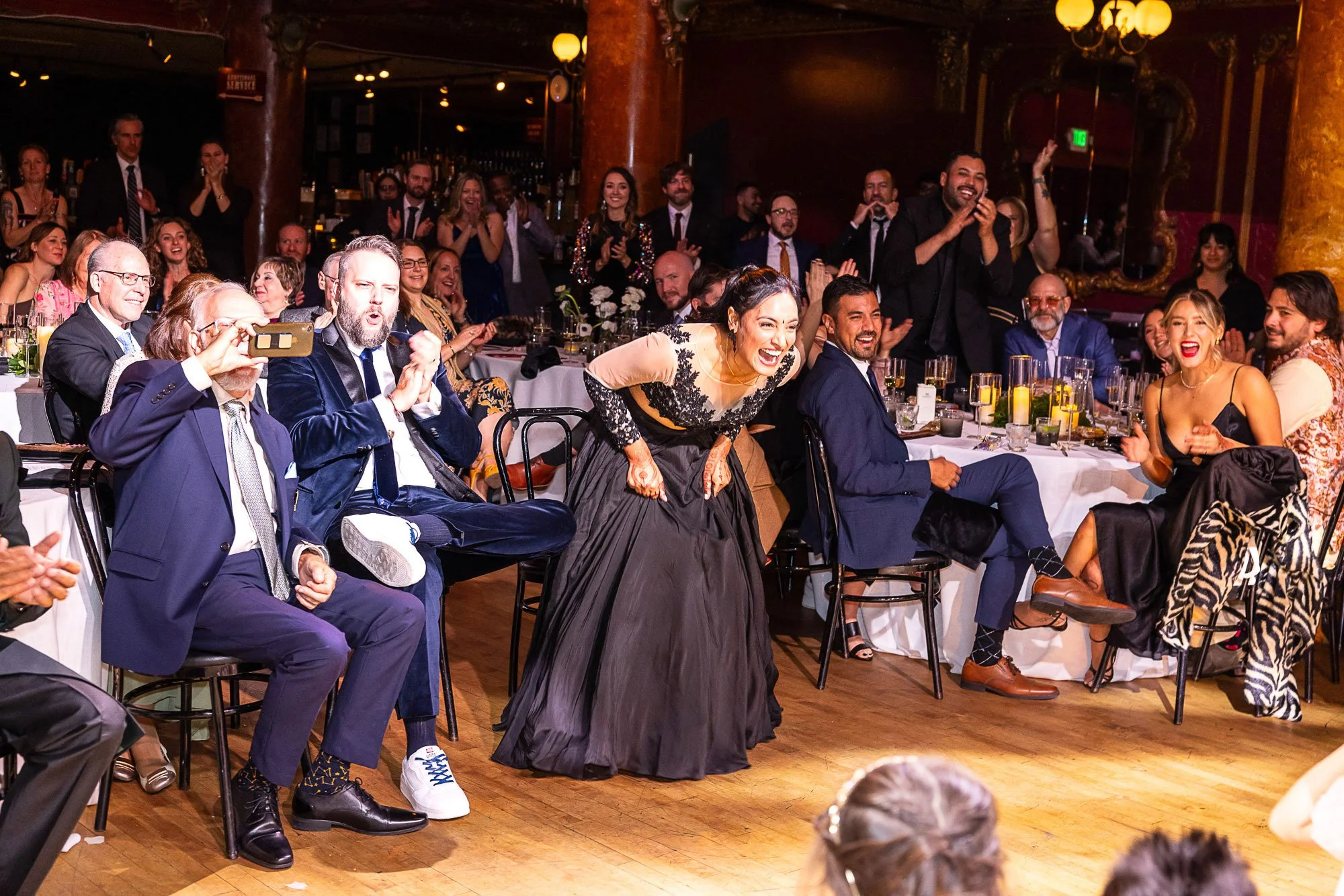 A lively indoor event with a diverse group of people sitting and standing, clapping, and smiling. The setting includes decorated tables, warm lighting, and ornate red and gold decor.
