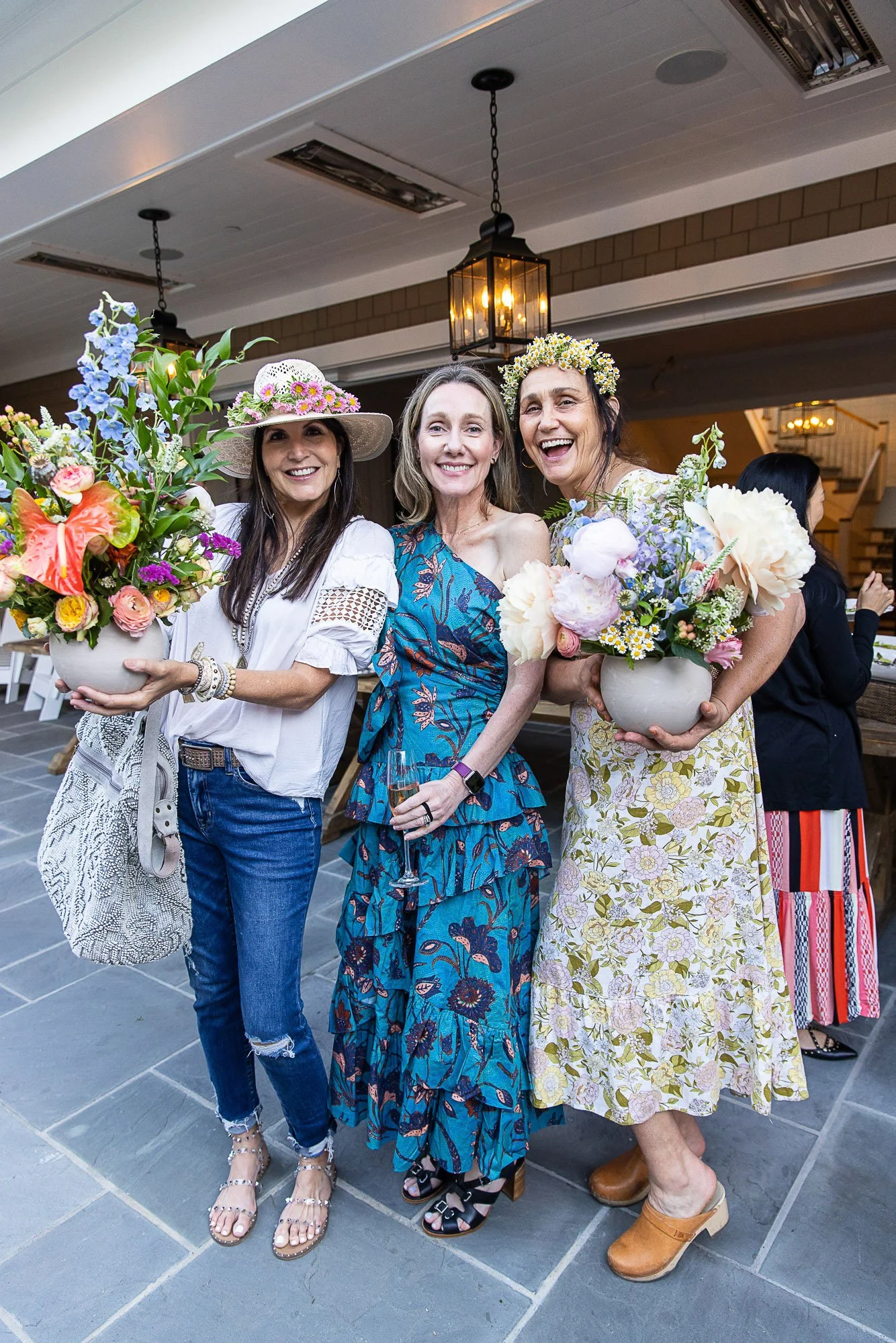 Three women in floral dresses holding flower arrangements pose together at an indoor event, smiling broadly.