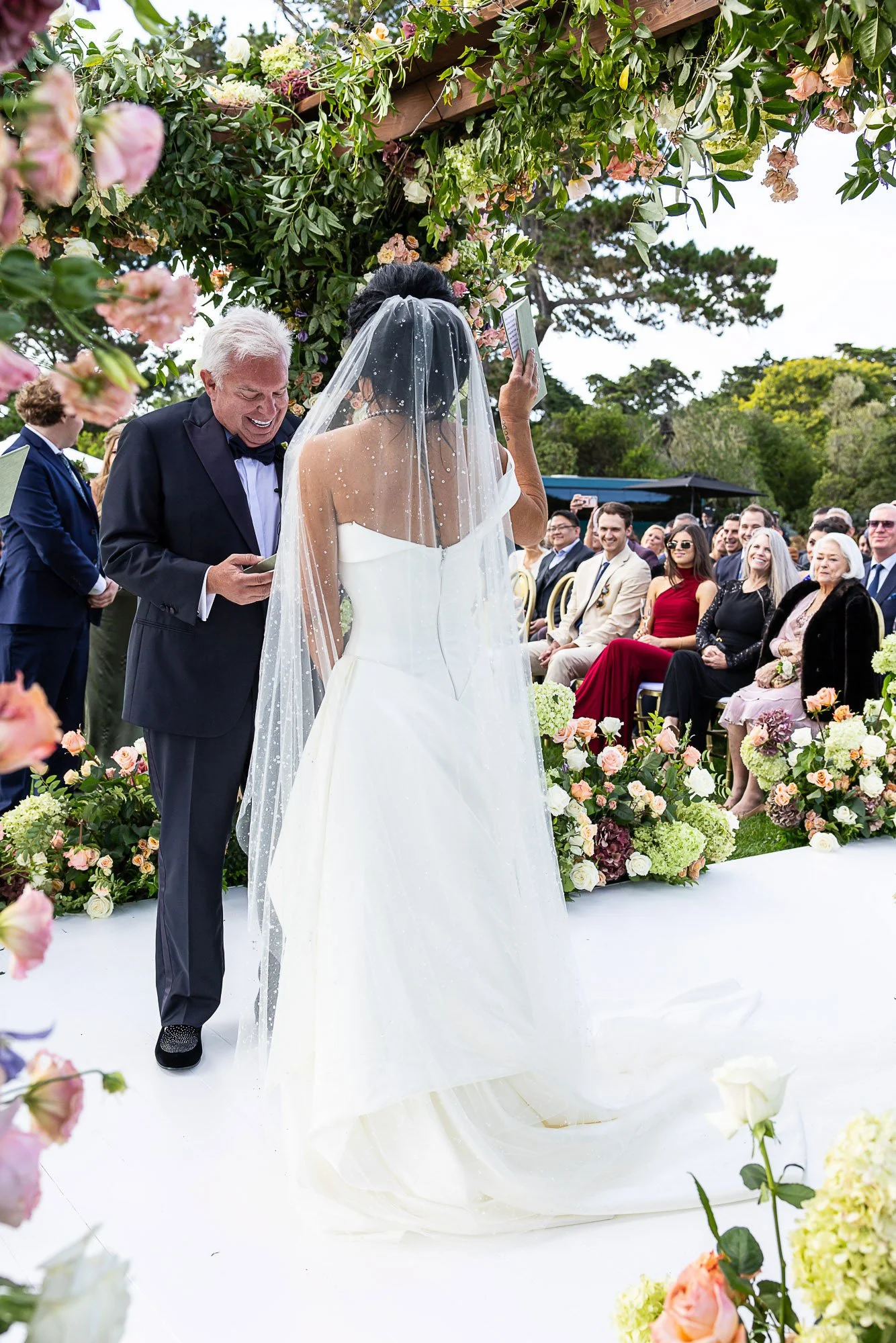 A bride and groom exchange vows at an outdoor wedding ceremony under a floral arch. The bride wears a white gown and veil, and the groom wears a black tuxedo. Guests sit and watch the ceremony, surrounded by colorful flowers and greenery.