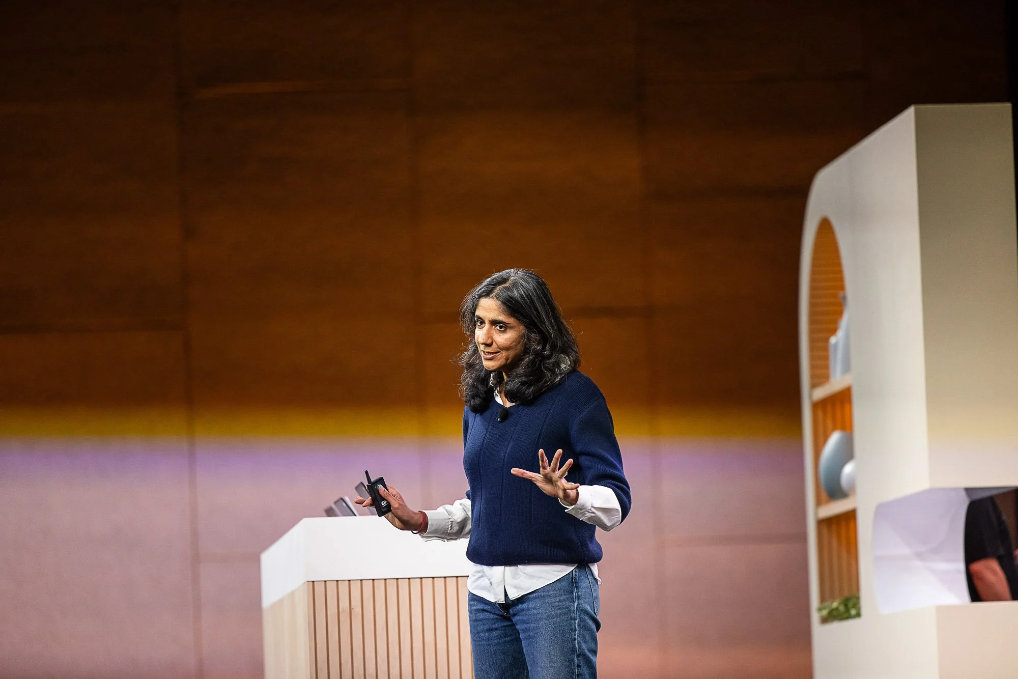 A woman with dark, wavy hair speaking at a presentation, gesturing with one hand while holding a device in the other, in a room with wooden-paneled walls and a white and wood podium.