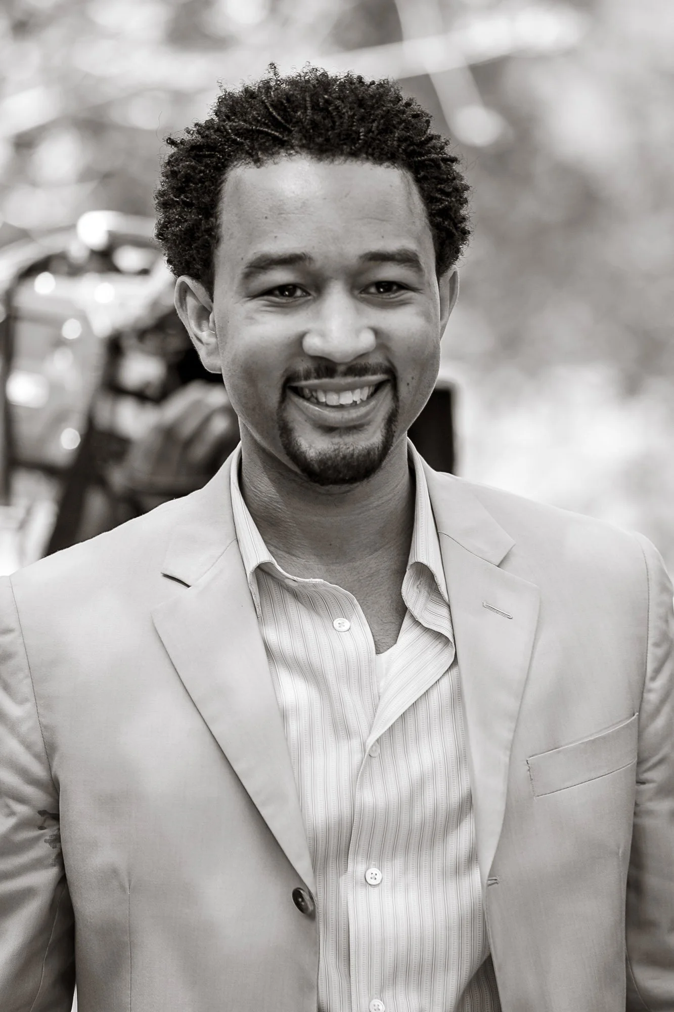 Black and white portrait of John Legend wearing a suit and dress shirt, standing outdoors with blurred background.