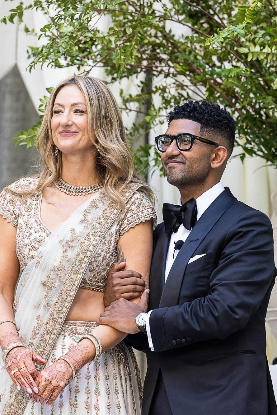 Bride in an ivory and gold embroidered lehenga with mehndi on her hands, arm in arm with the groom in a black tuxedo, at the Conservatory at One Sansome in San Francisco