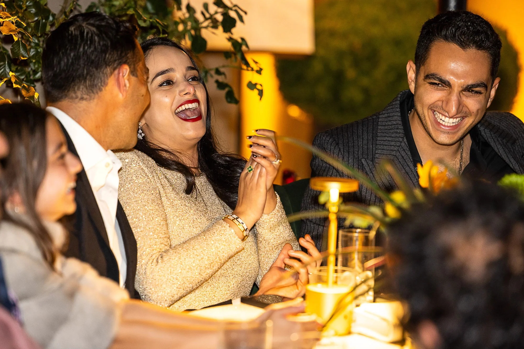People at a social gathering laughing and talking, with a focus on a woman wearing a gold dress and bright red lipstick.
