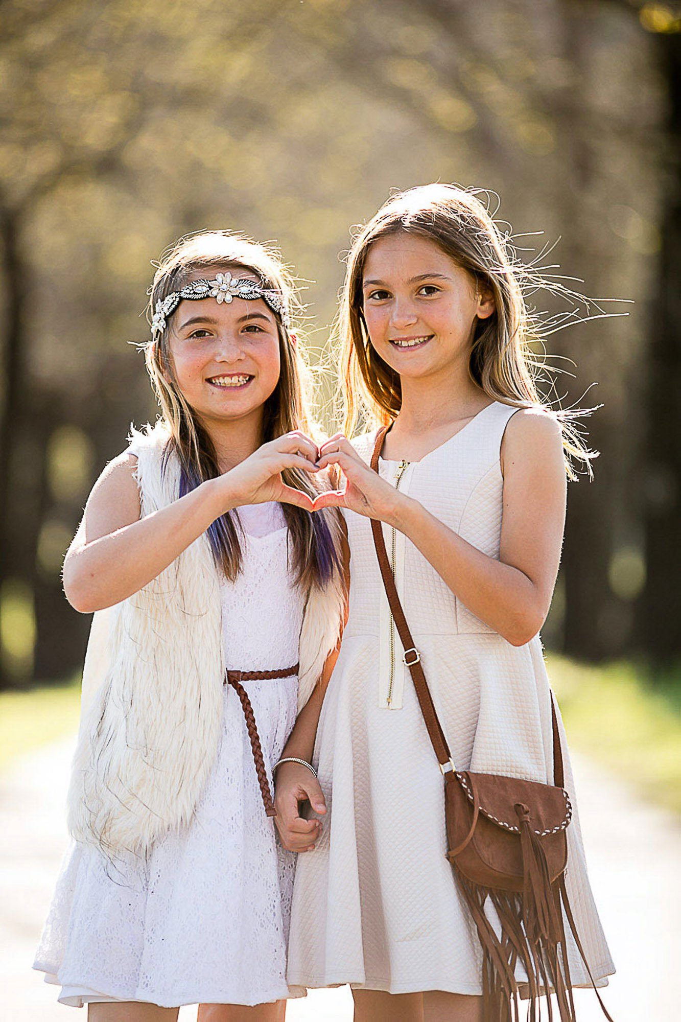 Two young girls smiling outdoors in a park, forming a heart shape with their hands.