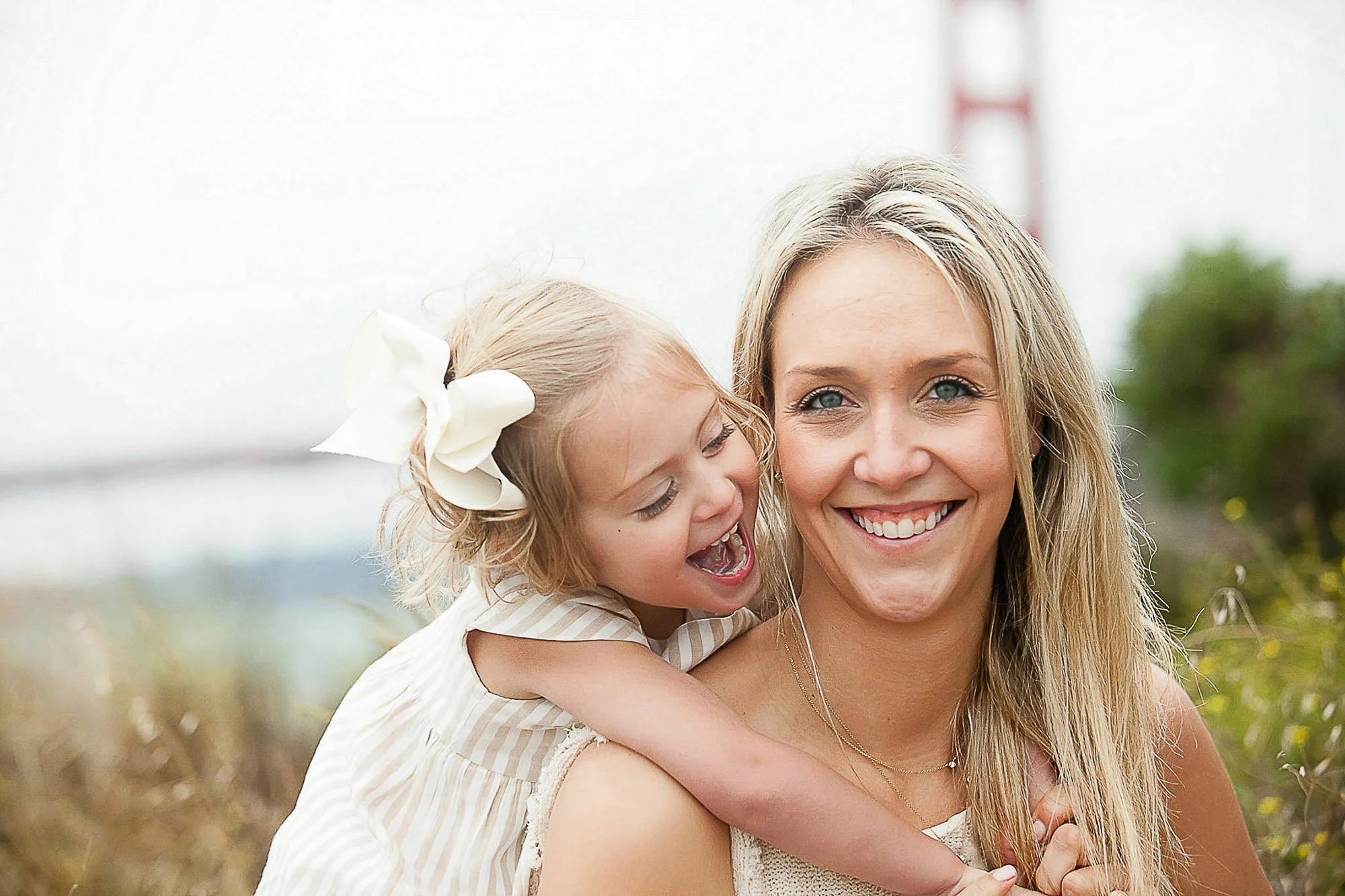 A woman smiling while holding a young girl on her back, at Travis Marina in Sausalito with the Golden Gate Bridge in the background.