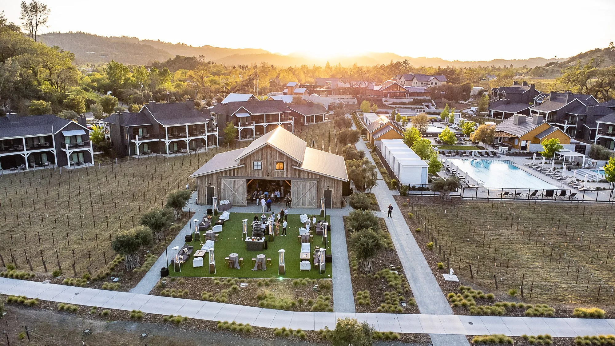 Aerial view of Napa Valley Four Seasons outdoor event space, old barn, pool, modern houses, and surrounding hills during sunset.