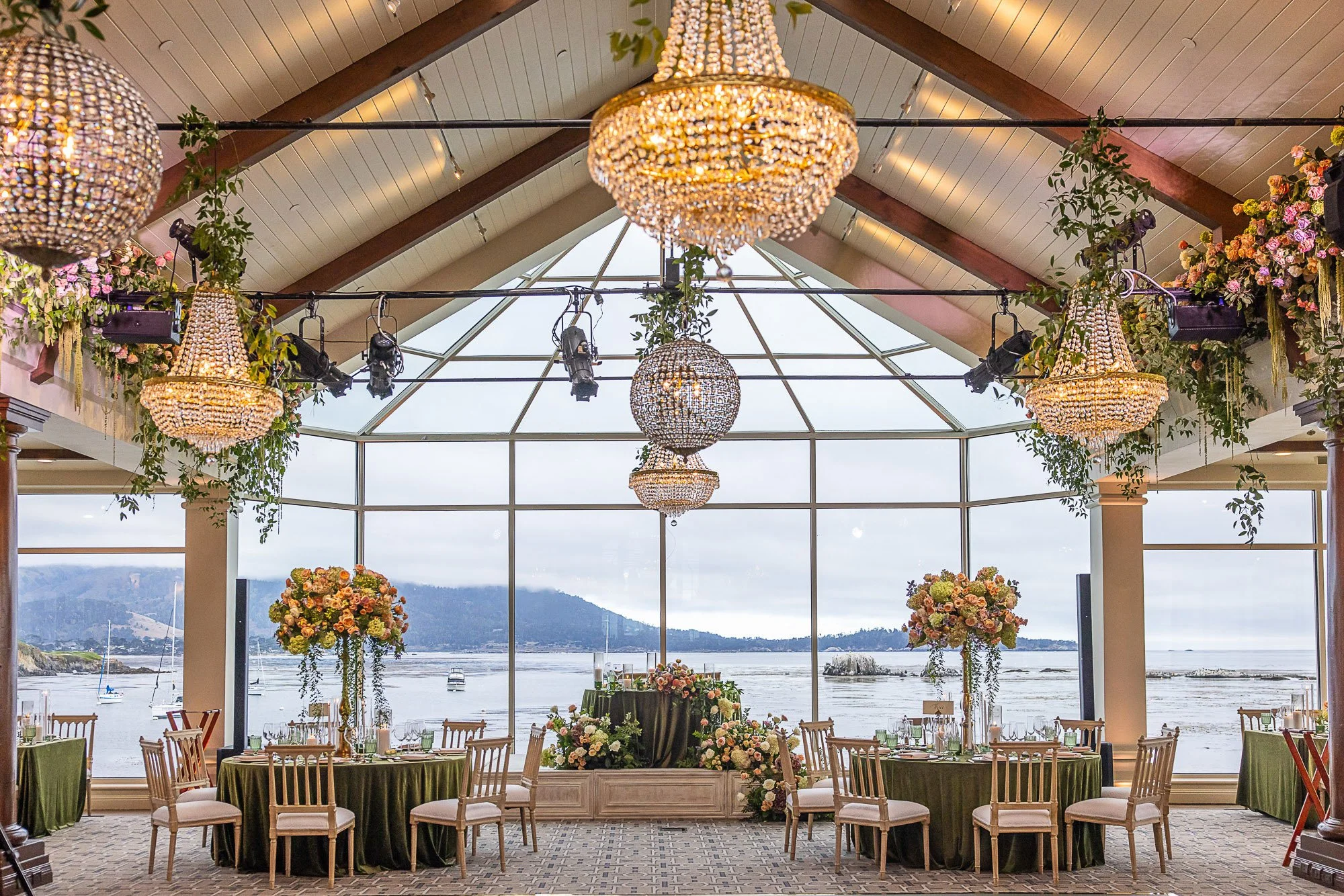 Elegant banquet reception area with round tables with green tablecloths, floral centerpieces, and chairs. Crystal chandeliers hanging from a high, wooden ceiling at the Beach and Tennis Club in Pebble Beach, CA.