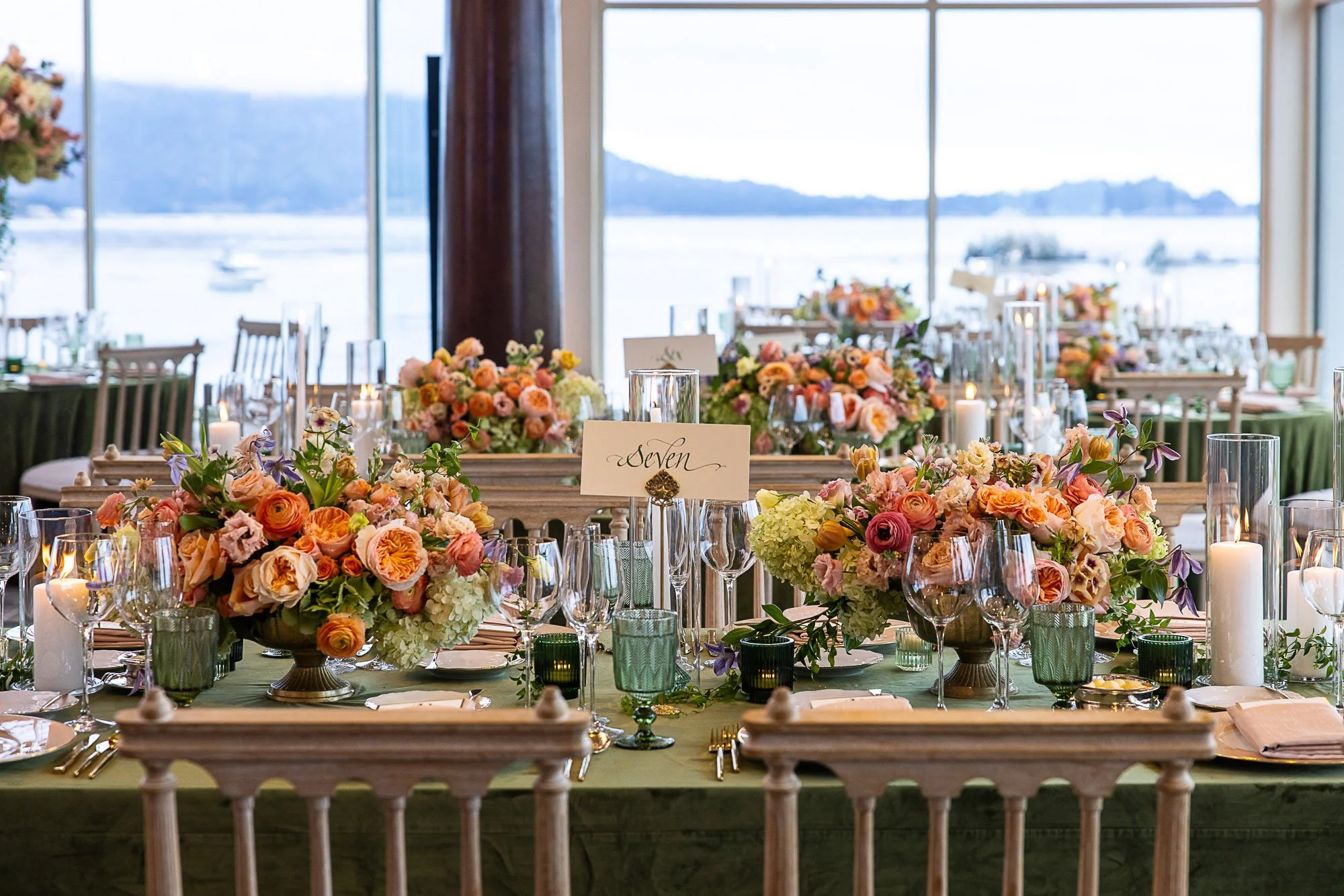 Elegant wedding reception table decorated with colorful flower arrangements, candles, and glassware, set near large windows with a view of a water body and mountainous landscape at the Beach and Tennis Club in Pebble Beach, CA.