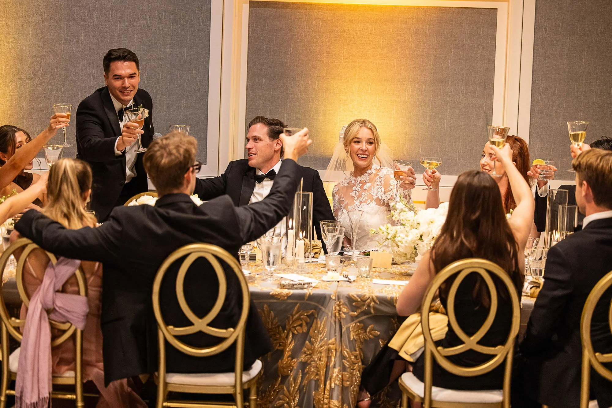 A wedding reception with guests in formal attire raising glasses for a toast around a decorated table, featuring a smiling bride and groom, festive lighting, and elegant gold and black chairs at Cavallo Point in Sausalito, CA.