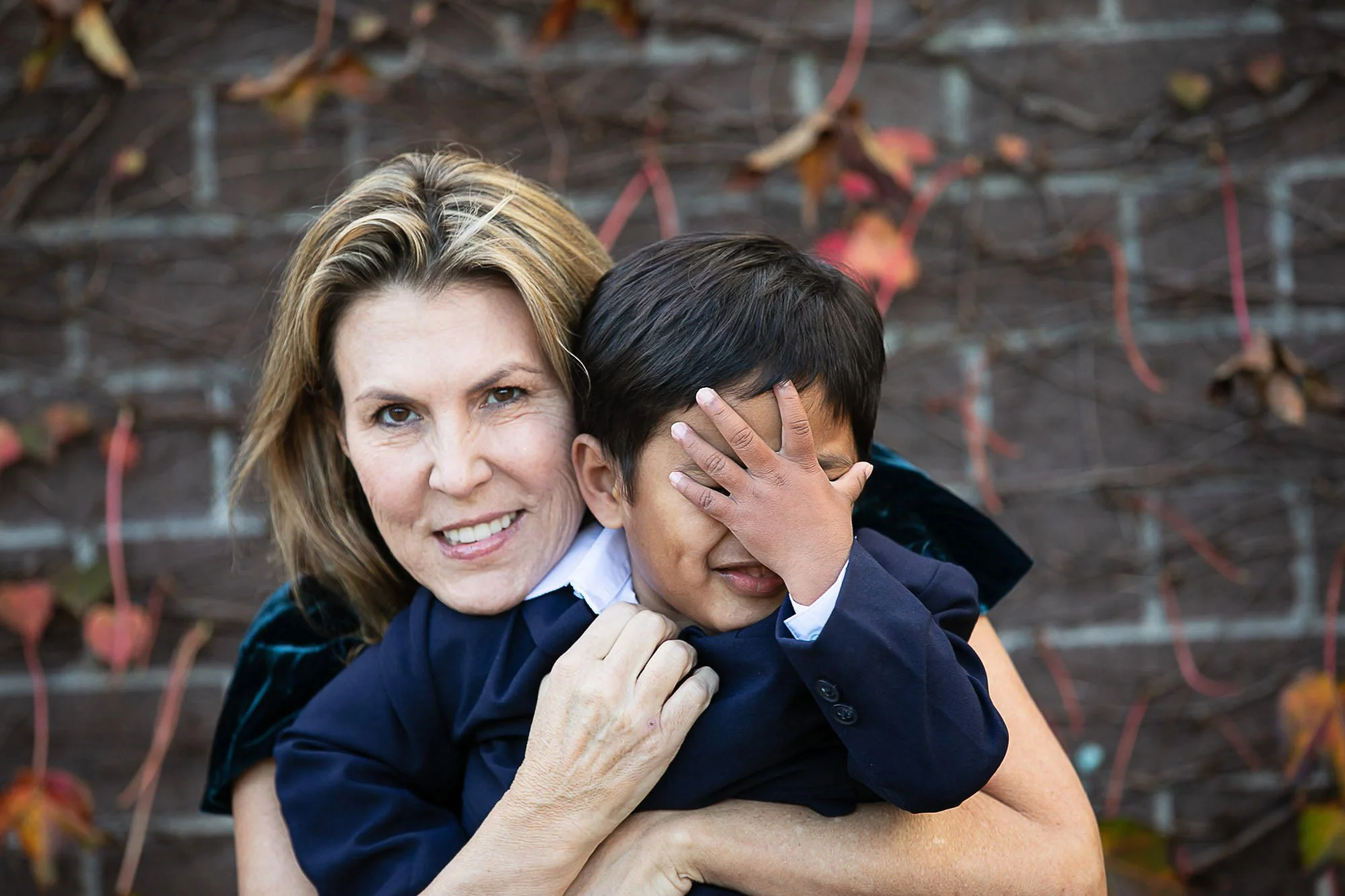 A woman with blonde hair embracing a young boy with dark hair. They are smiling and holding each other outdoors in front of a vine-covered wall with autumn leaves.