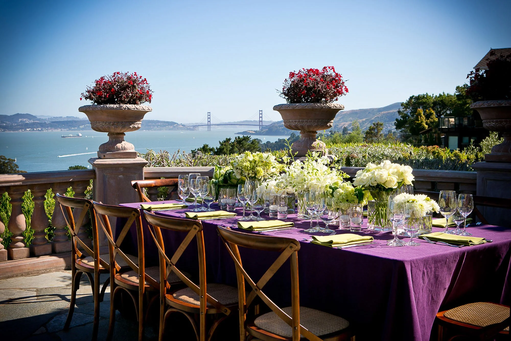A dining table set outdoors with a purple tablecloth, glassware, and floral centerpieces, overlooking the water with a bridge in the background.