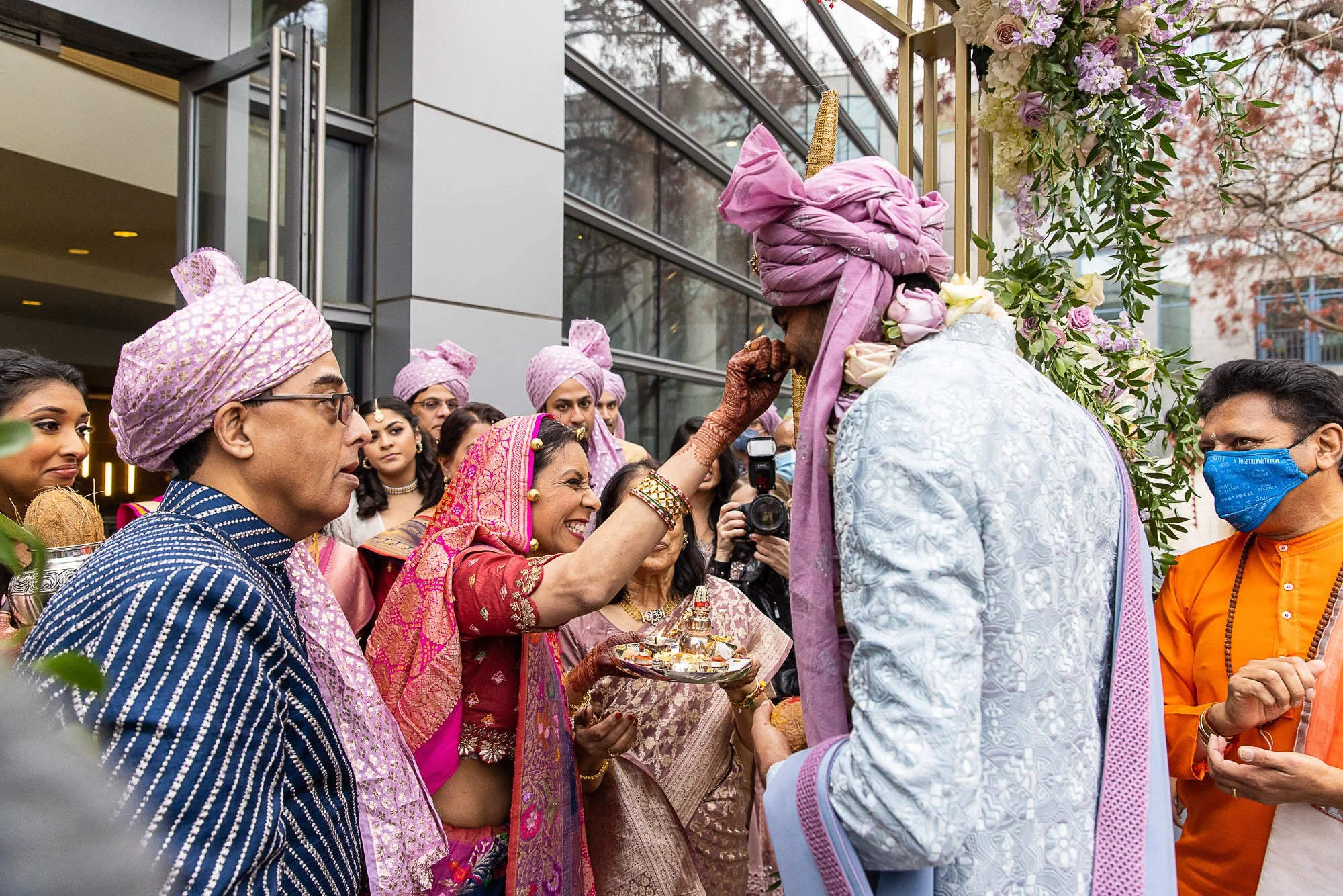A group of people dressed in traditional Indian attire participating in a ceremony outdoors, with a woman applying a red mark on a man's forehead who is wearing a pink turban, surrounded by onlookers and floral decorations.