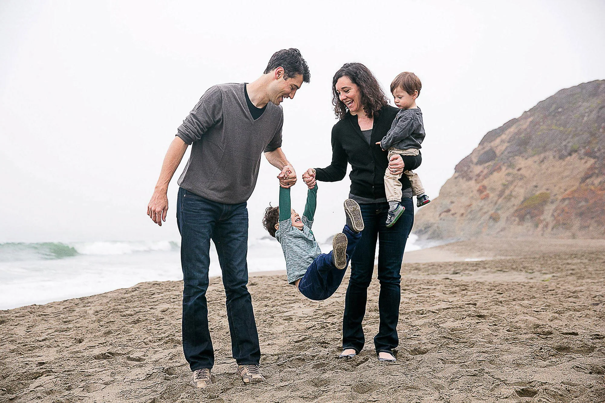 A family of four enjoying time on the beach. The father, mother, and two children are smiling and holding hands. The mother is holding one child while the father is holding the other child's hands, swinging him playfully in the air on the sand near t