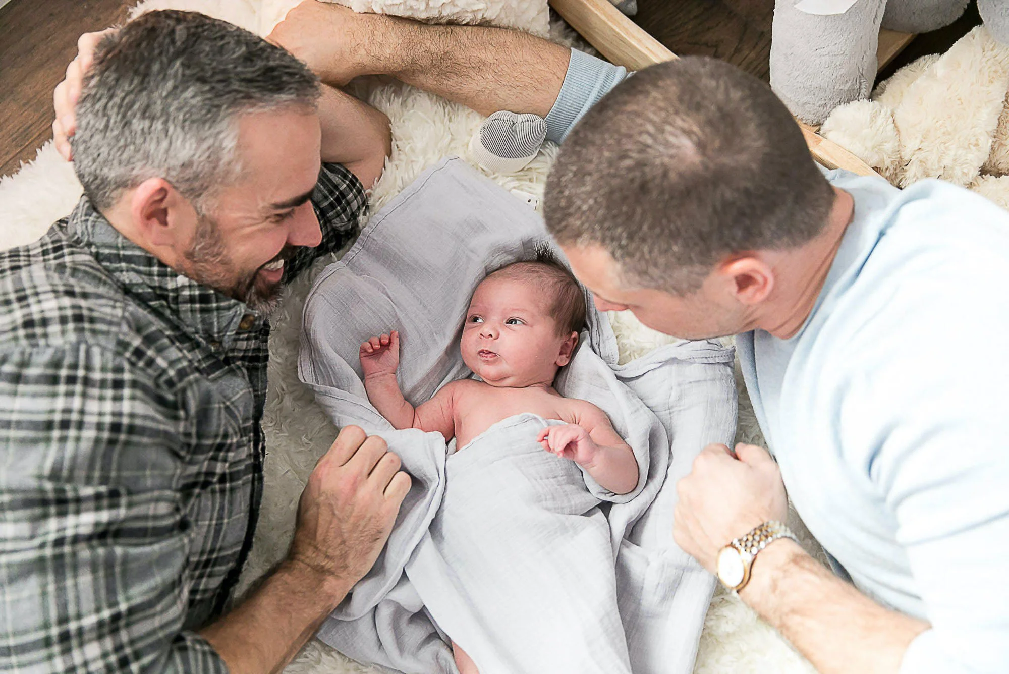 Two men lying on the floor, smiling and looking at a baby wrapped in a blanket, surrounded by plush rugs and cushions.
