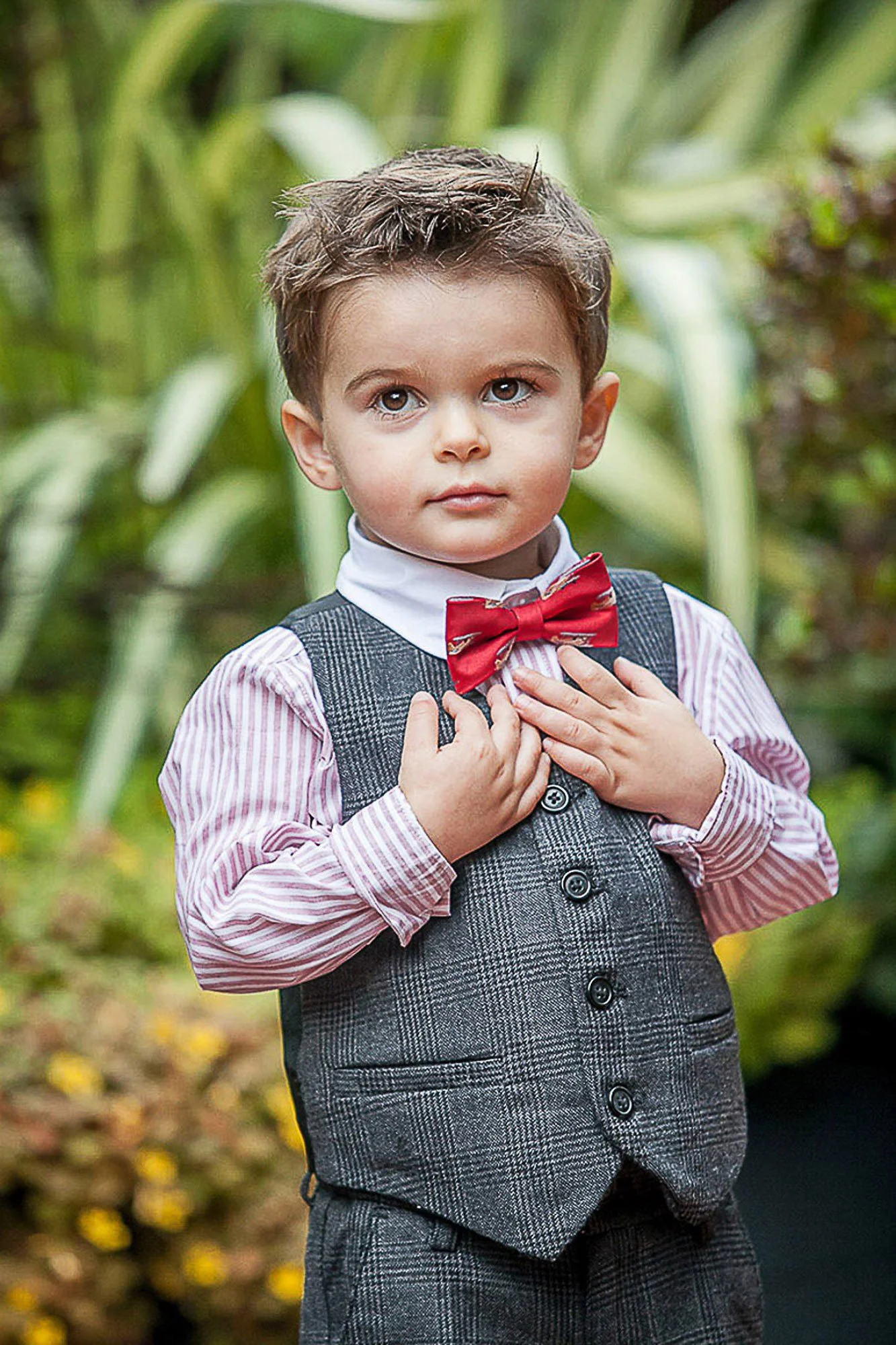 A young boy with brown hair and light skin is dressed in vintage-style clothing, including a gray checkered vest, white shirt with pink stripes, and a red bow tie. He stands outdoors in front of green foliage, with his hands on his chest, looking dir