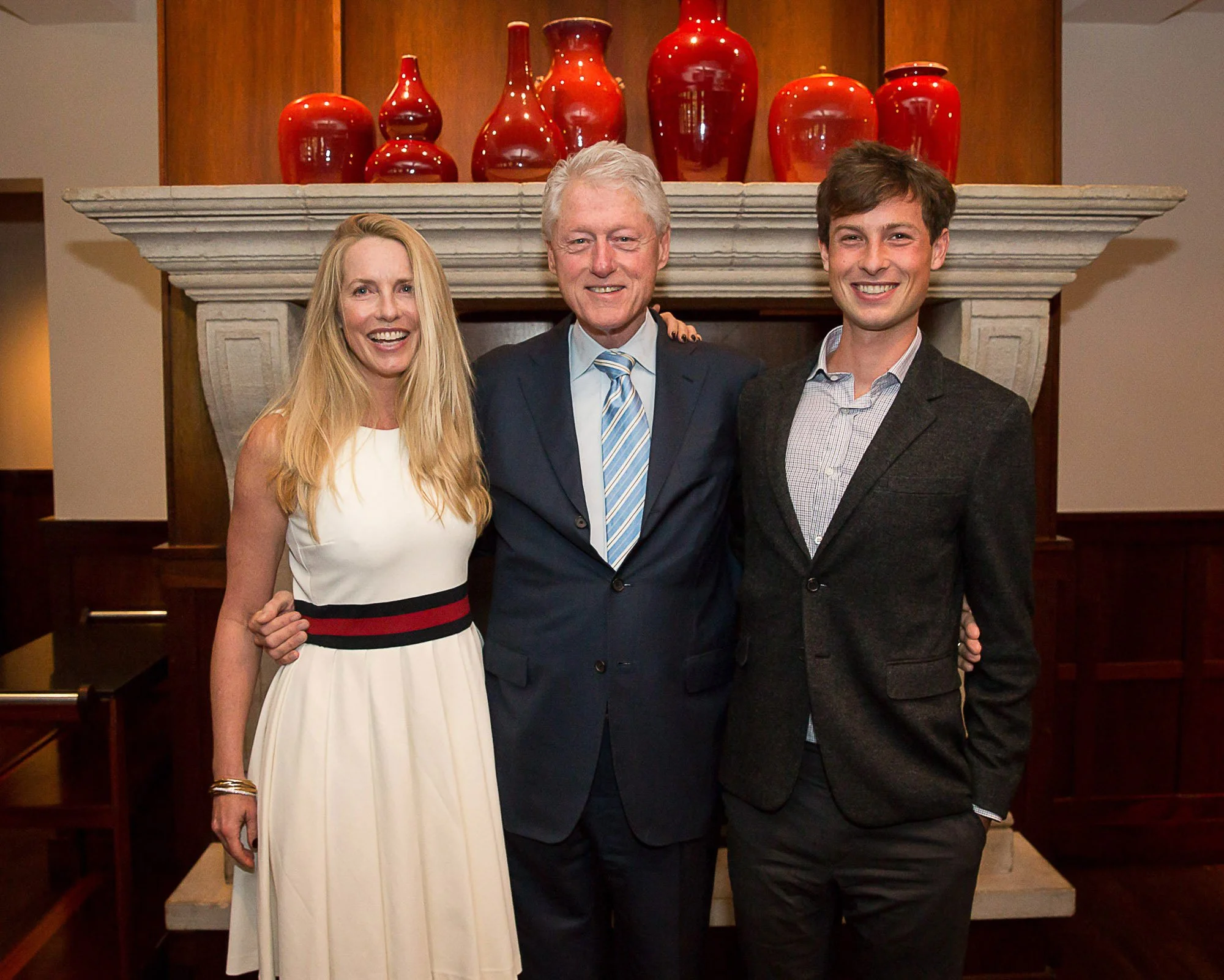 Bill Clinton standing in front of a fireplace with red vases on the mantel. The woman on the left has long blonde hair and is wearing a white dress. The man in the middle has gray hair and is dressed in a dark suit with a light blue tie. 
