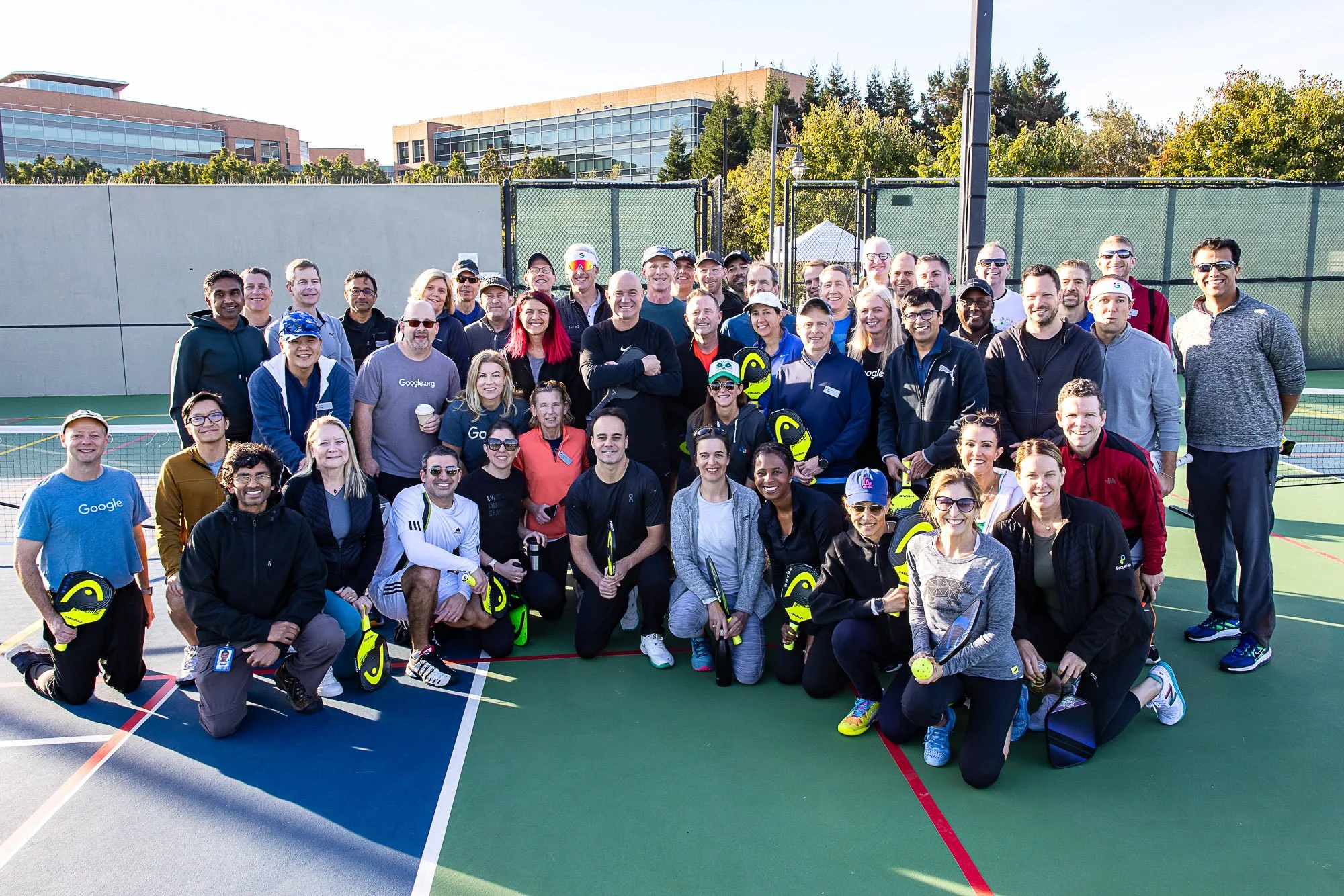 Group of people gathered on a tennis court, holding tennis rackets and wearing casual and sportswear, smiling at the camera during daytime.