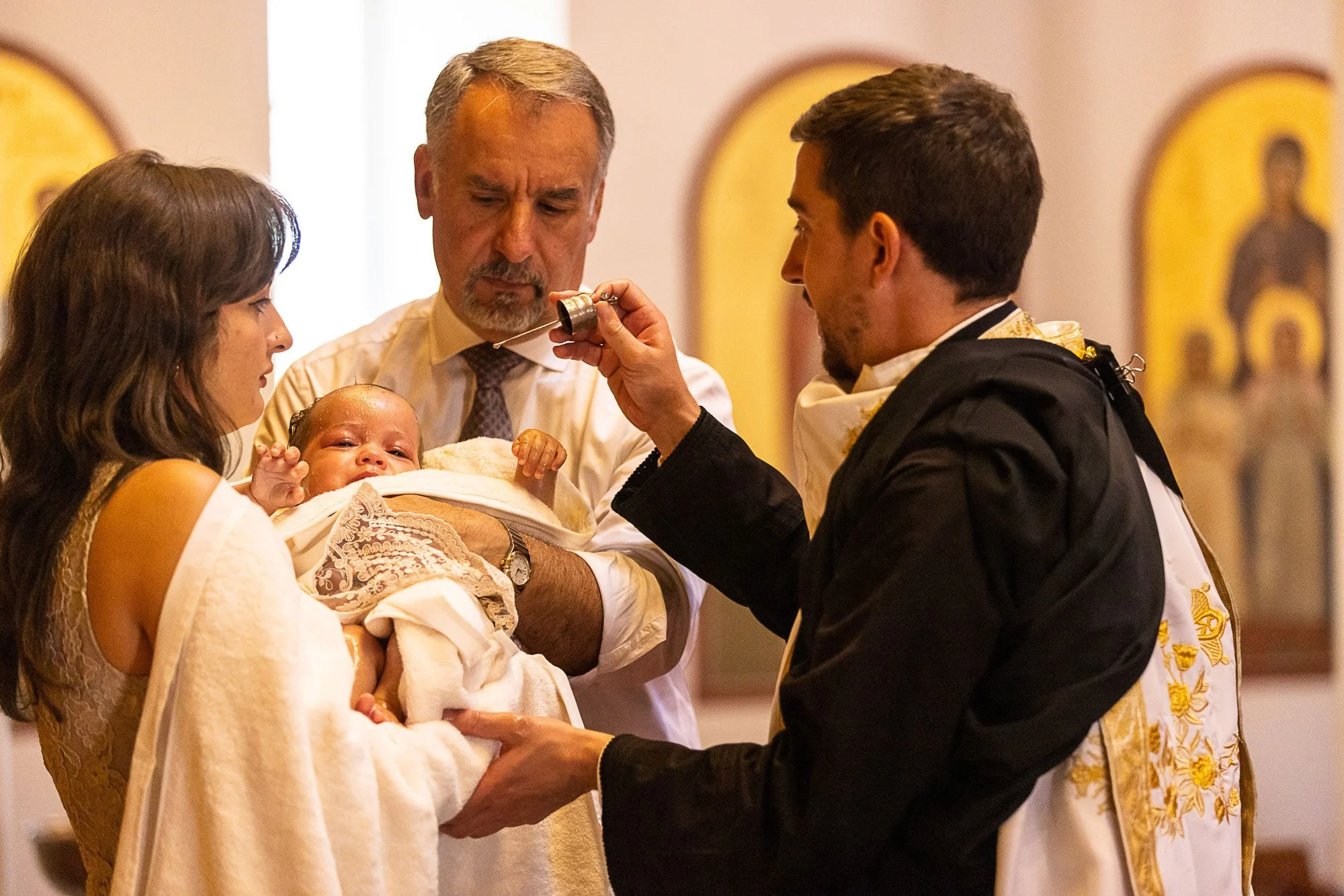 A baptism ceremony with a priest performing the baptism, a woman holding a baby, a man assisting with the ceremony, and religious icons in the background.