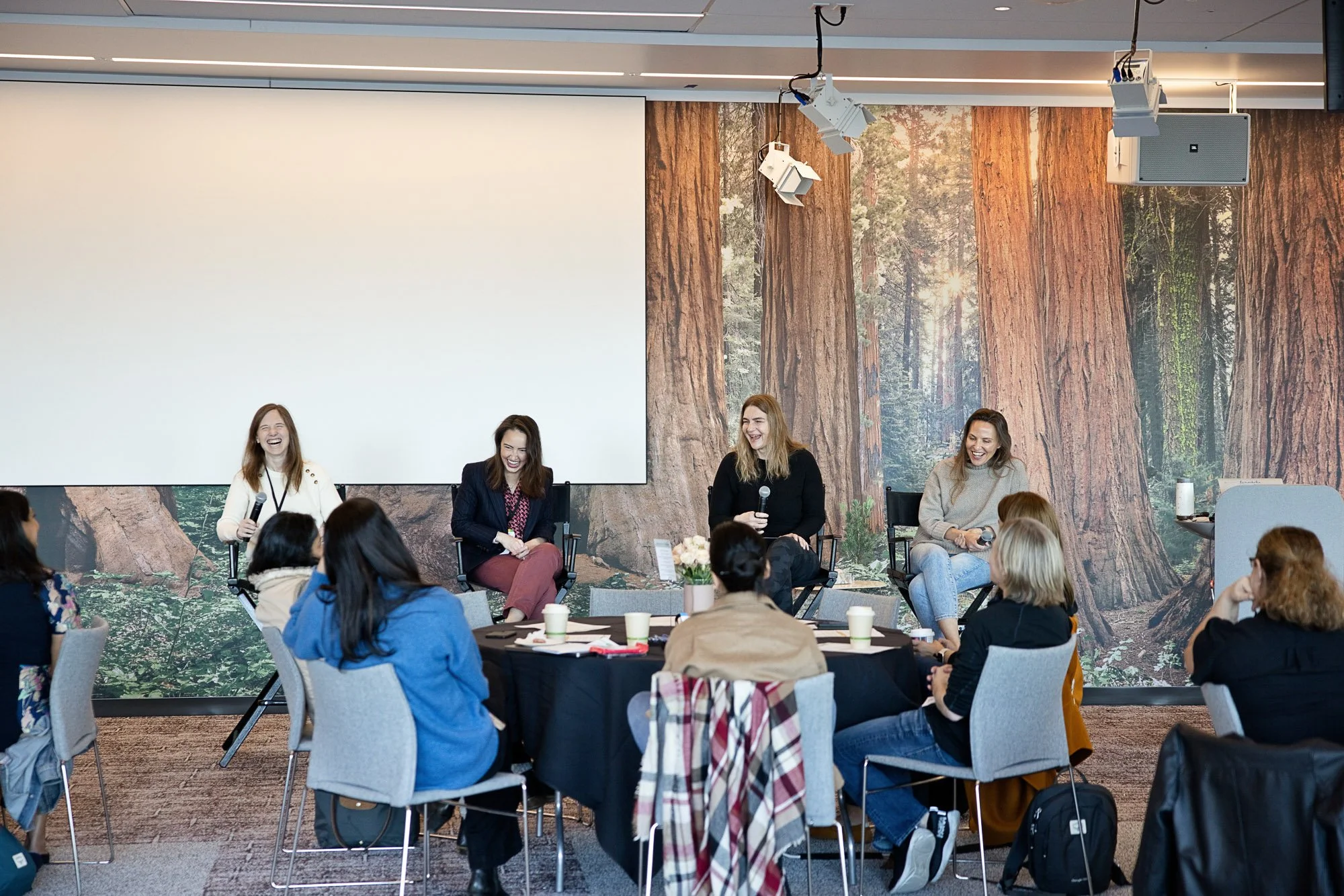 Four women sitting on a panel in front of an audience in a conference room with a forest-themed backdrop. The women are holding microphones and smiling, with attendees seated at round tables listening.