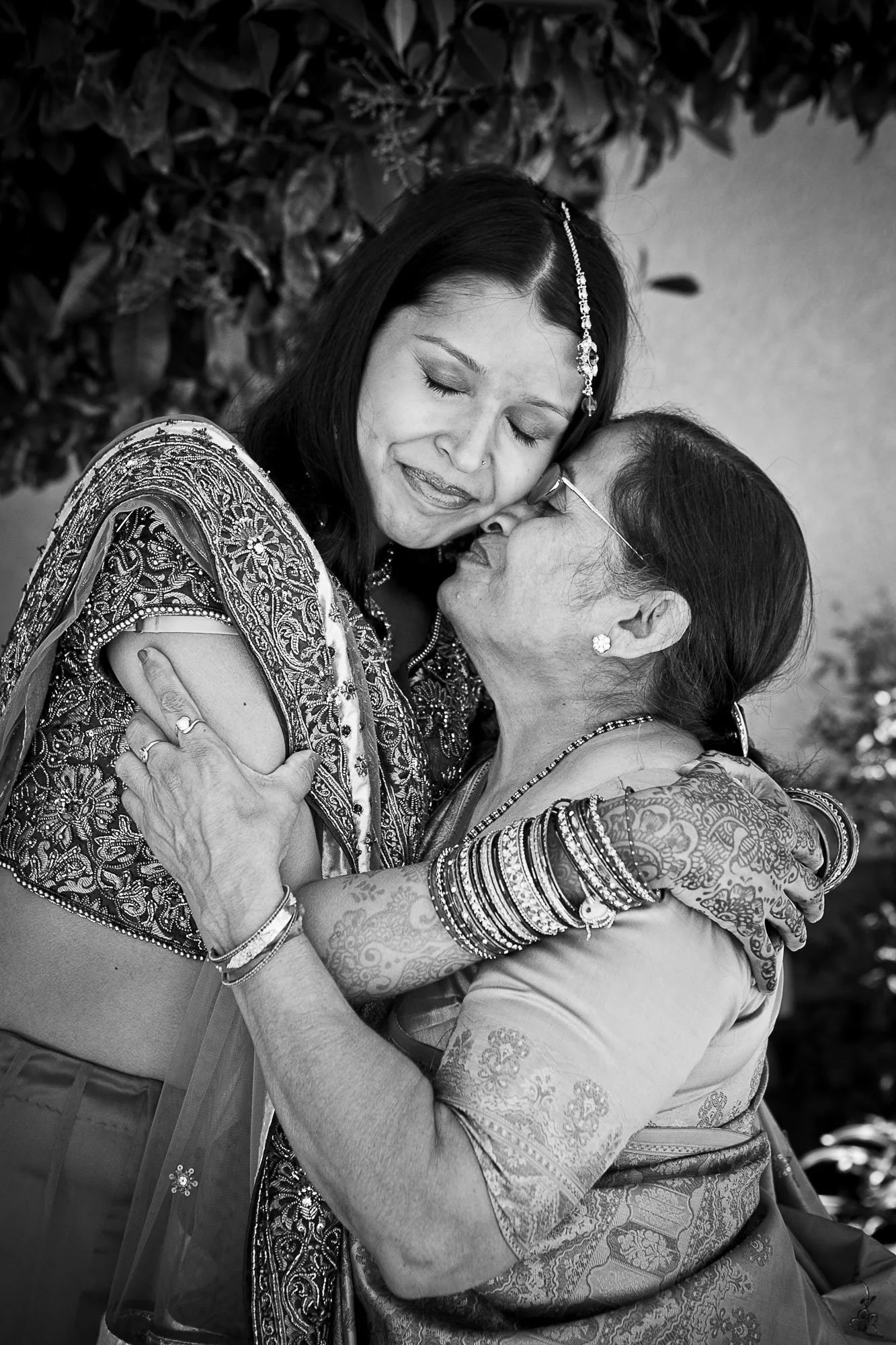 Black and white photo of two women embracing, eyes closed, wearing traditional Indian clothing and jewelry, outdoors with trees in the background.