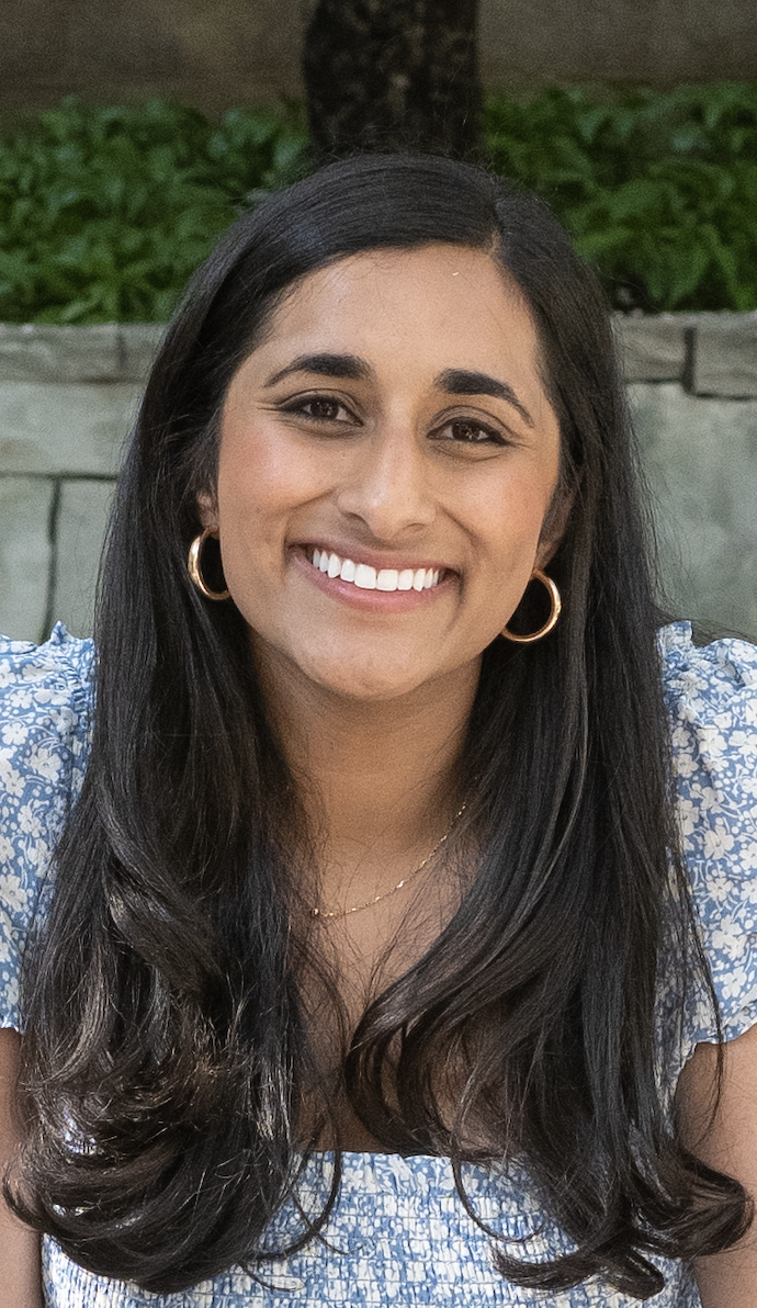 A woman with long dark hair, wearing gold hoop earrings and a blue floral dress, smiling outdoors with greenery in the background.