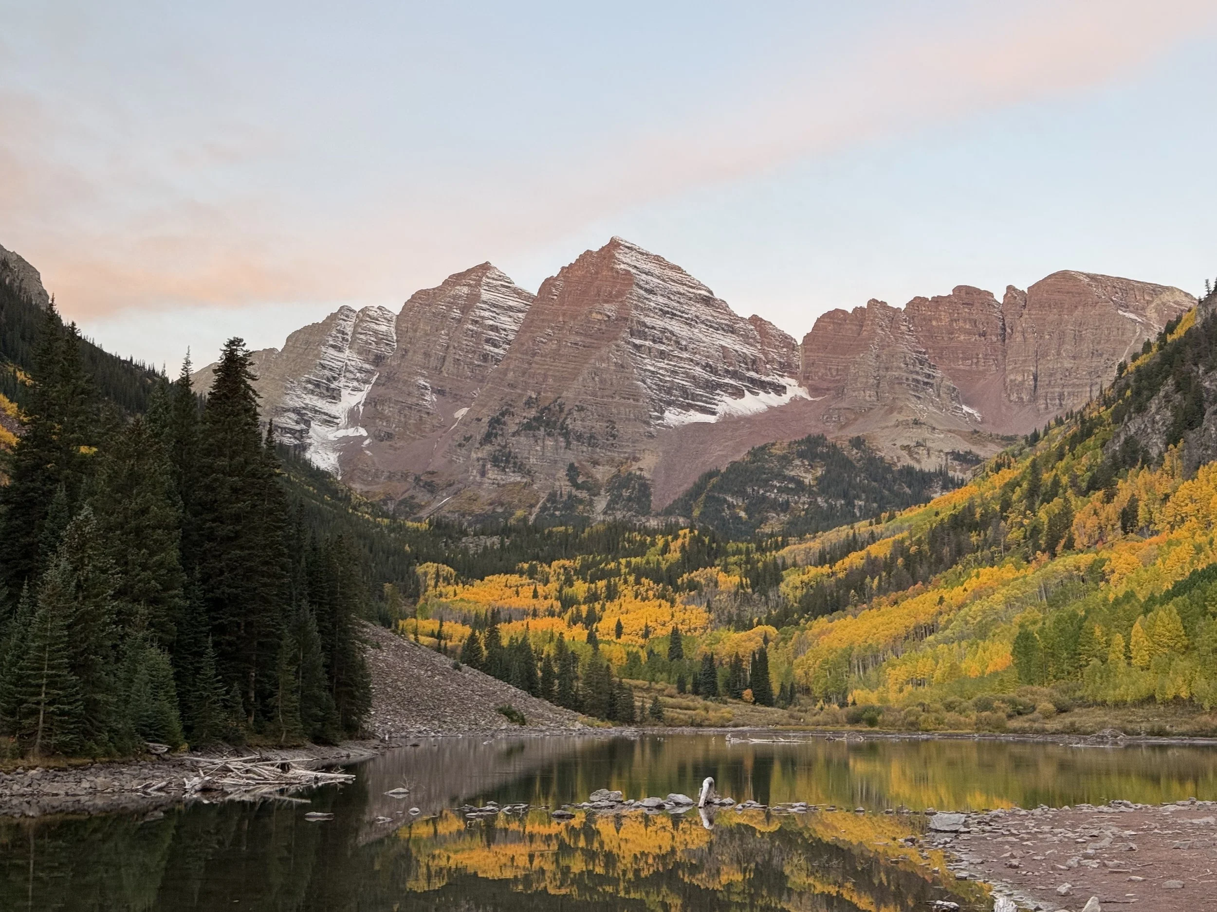 Snow-capped mountain peaks with forested slopes and colorful fall foliage near a calm river at sunset.