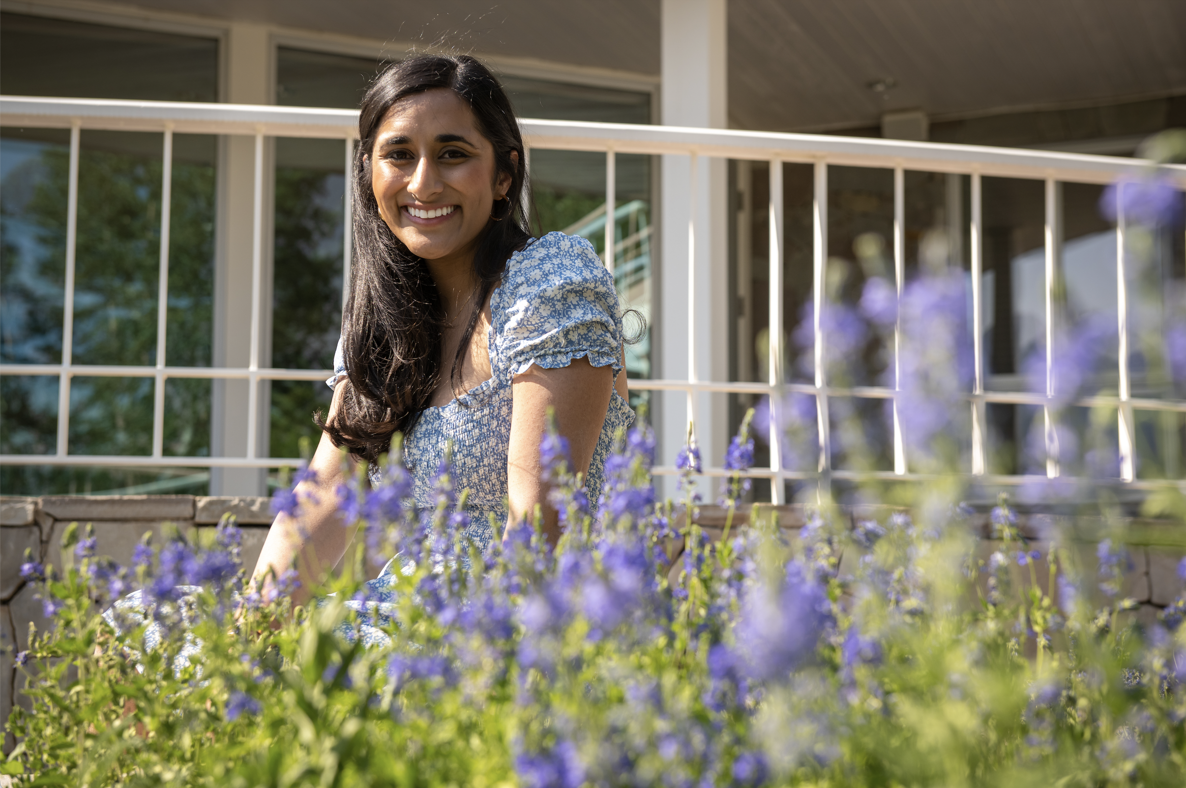 A young woman with long dark hair smiling outdoors, wearing a blue floral dress, sitting by purple flowers in a garden with a house and balcony in the background on a sunny day.