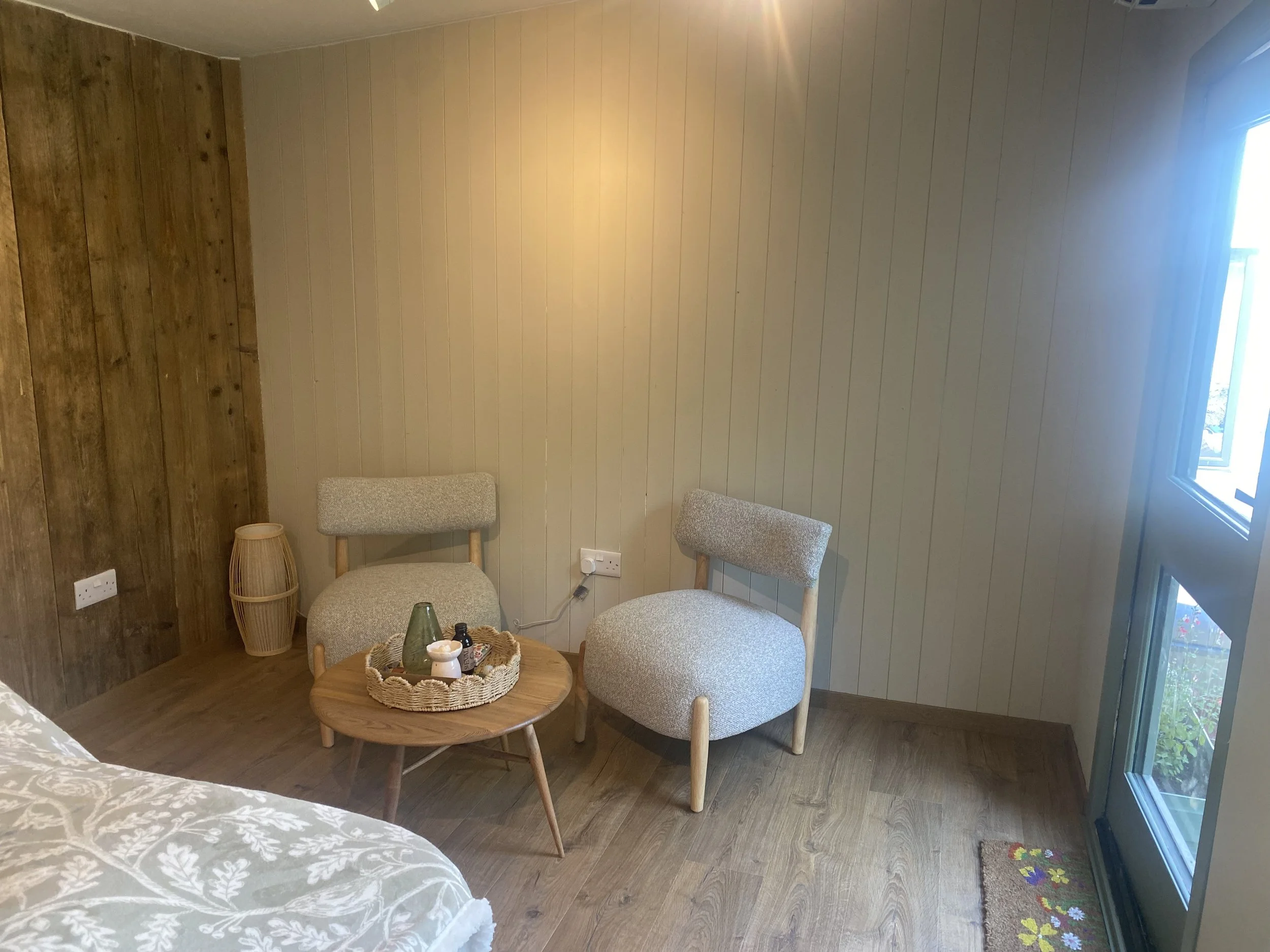 A cozy corner of a room with wooden and beige panel walls, two upholstered chairs around a small round wooden table holding a basket with decorative items, near a window with curtains, and a small colorful floral rug on the floor.