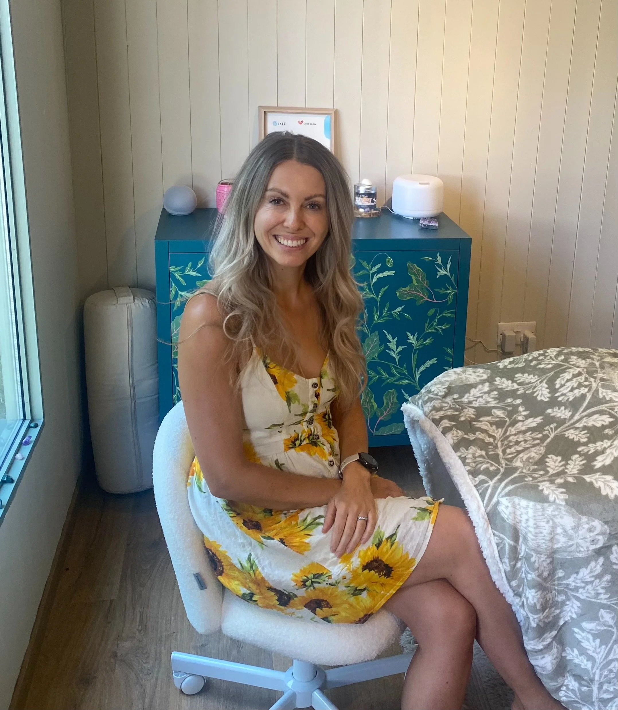 A woman smiling while sitting on an office chair in a room with beige paneled walls. She is wearing a yellow and white floral dress. Behind her, there is a blue cabinet with a painted leaf pattern, a framed certificate or sign, and various objects including a small speaker, a candle, and a humidifier. To the right, part of a bed with a floral bedspread and a power outlet with plugged-in cords are visible.