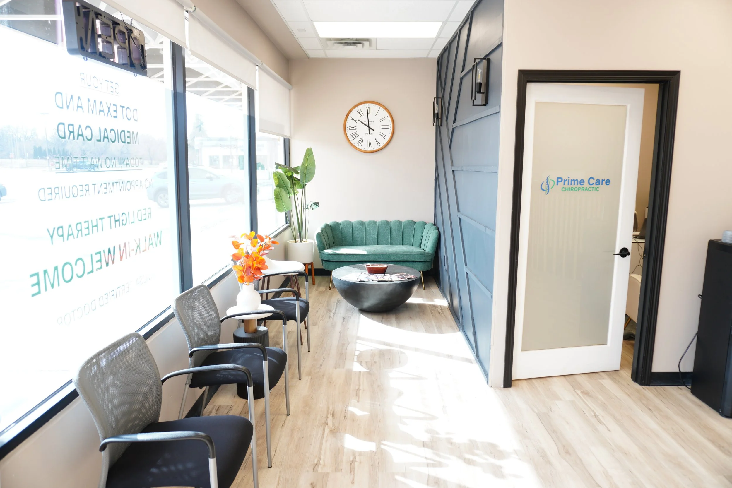 The image shows a waiting area inside a chiropractic office with a row of six black chairs along the window, a white round table with a vase of orange flowers, a green upholstered chair with a large potted plant behind it, a wall clock, and a door with a sign that says "Prime Care Chiropractic."