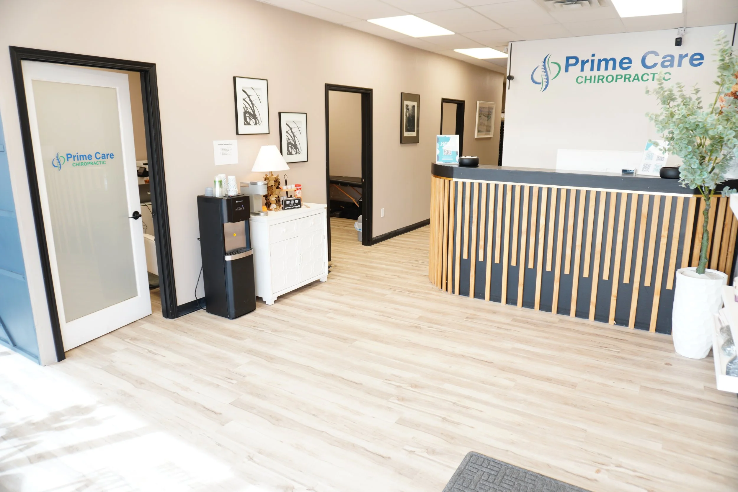 Reception area with a curved wooden counter, framed artwork on the wall, a water dispenser, a small white cabinet, and a glass door with the Prime Care Chiropractic logo.