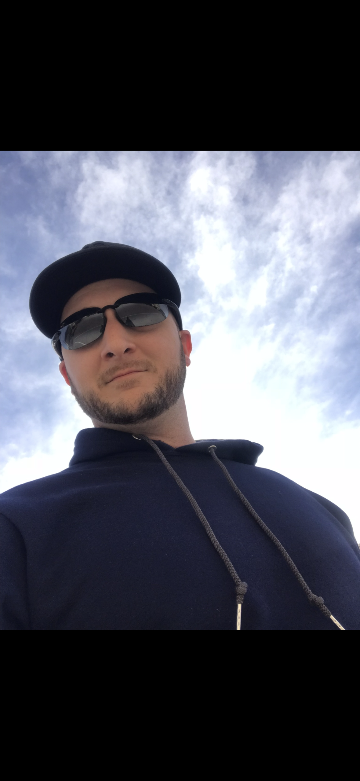 A man wearing sunglasses and a black hoodie, looking at the camera with a slight smile, outdoors with a cloudy sky in the background.