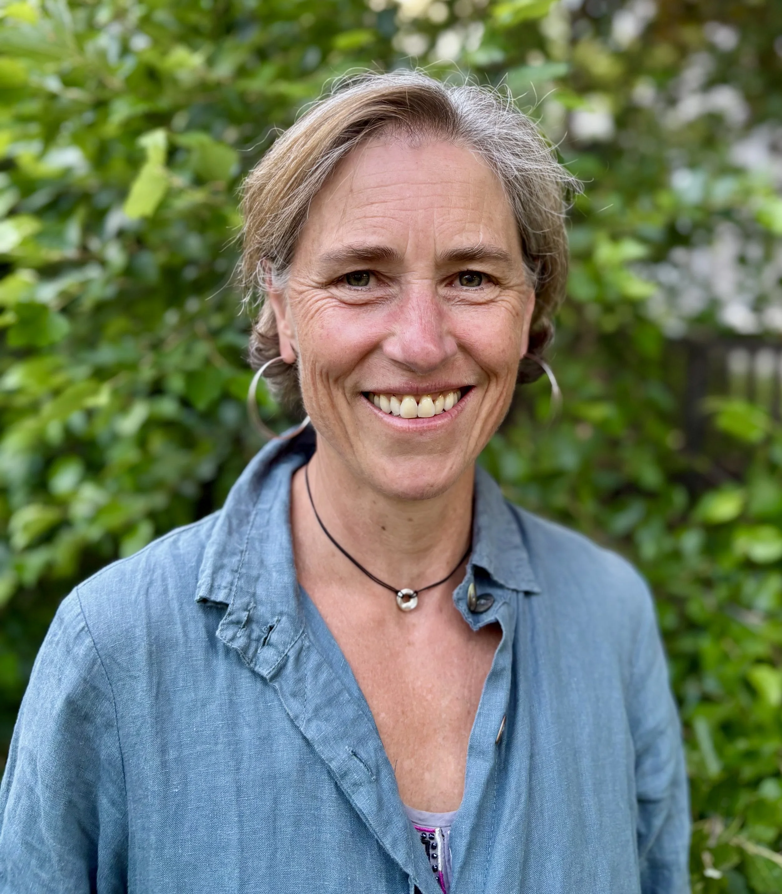 Close-up of a smiling middle-aged woman with short gray hair, wearing a blue denim shirt and hoop earrings, standing outdoors with blurred green foliage in the background.