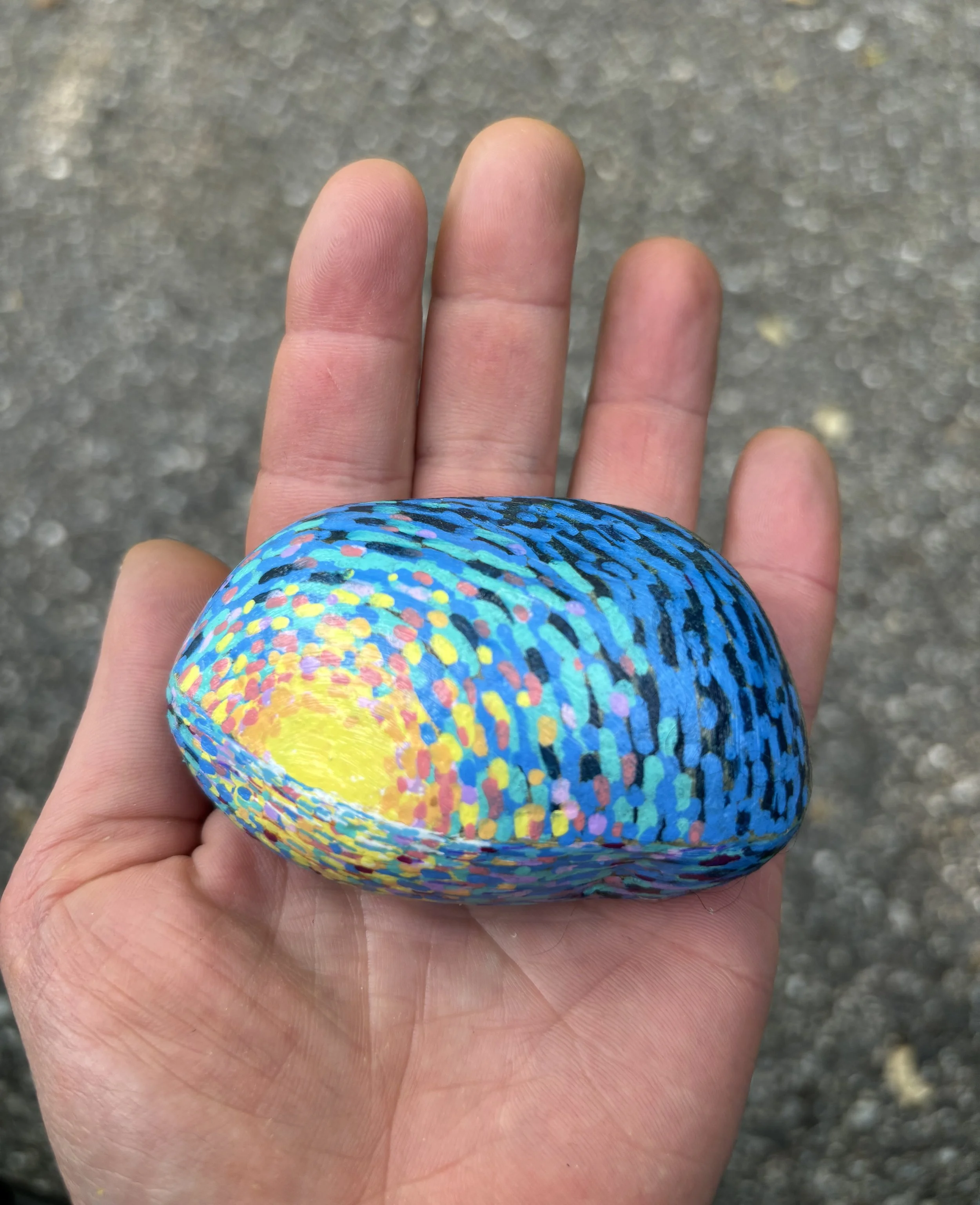 Close-up of a person's hand holding a colorful painted rock with a speckled pattern of blue, yellow, pink, black, and other colors, against a gray, gravelly background.
