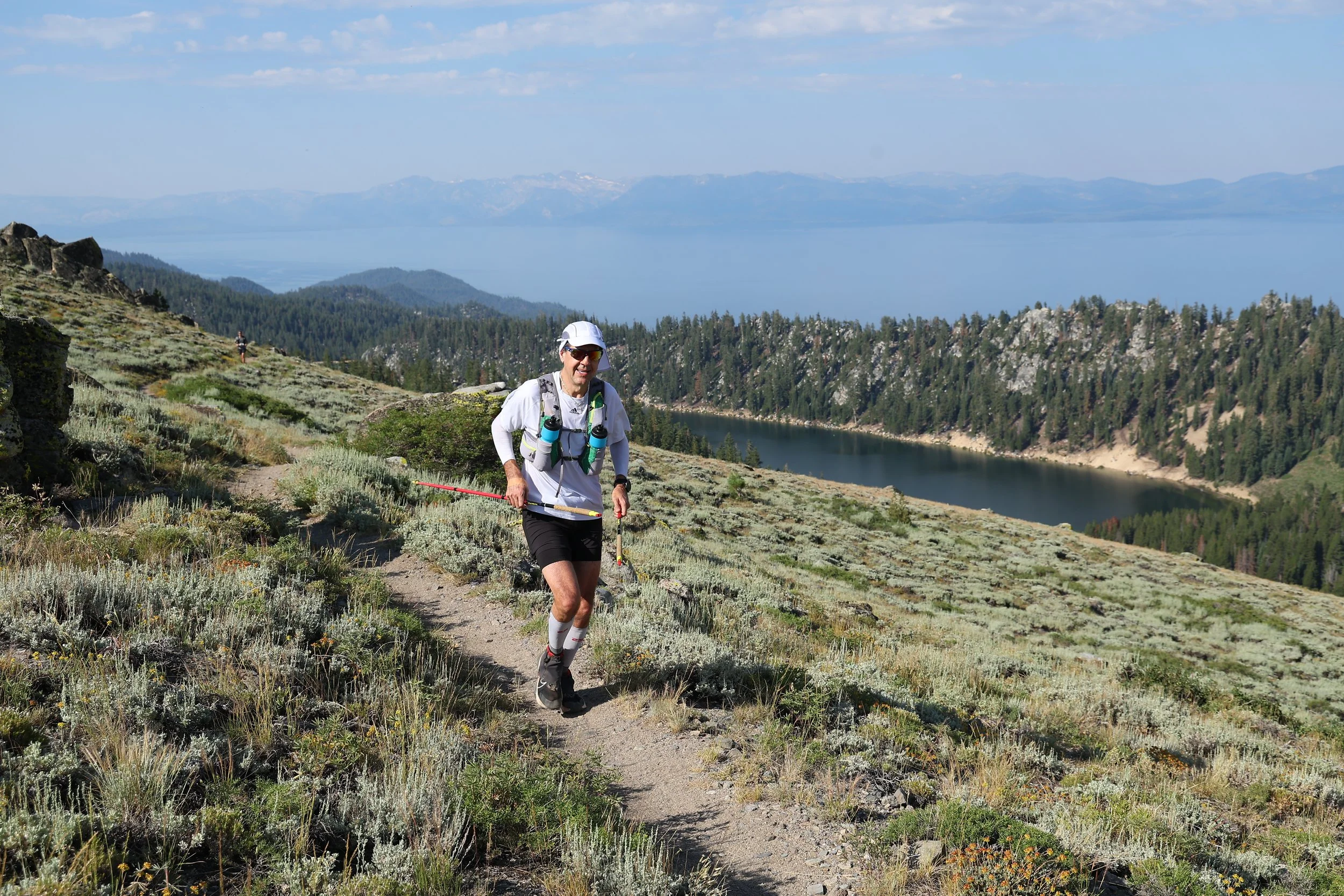 A man smiling and hiking on a trail in a mountainous area with a lake and forested hills in the background in lake tahoe with marlette lake in the near background.