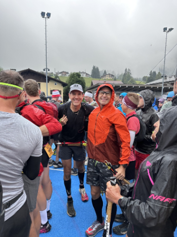 Two smiling runners with running gear and medals, surrounded by a crowd of runners in athletic clothing, under a cloudy sky at an outdoor skyrunning event.