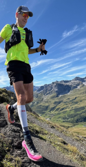 Male trail runner in bright yellow shirt, black shorts, pink running shoes, and sunglasses, running on a mountain trail with scenic green hills and mountains under a clear blue sky.  In the Alps