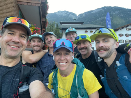 Group of seven smiling people in outdoor mountain setting, dressed in hiking gear with backpacks and caps, taking a selfie.  Transalpine run in the alps.