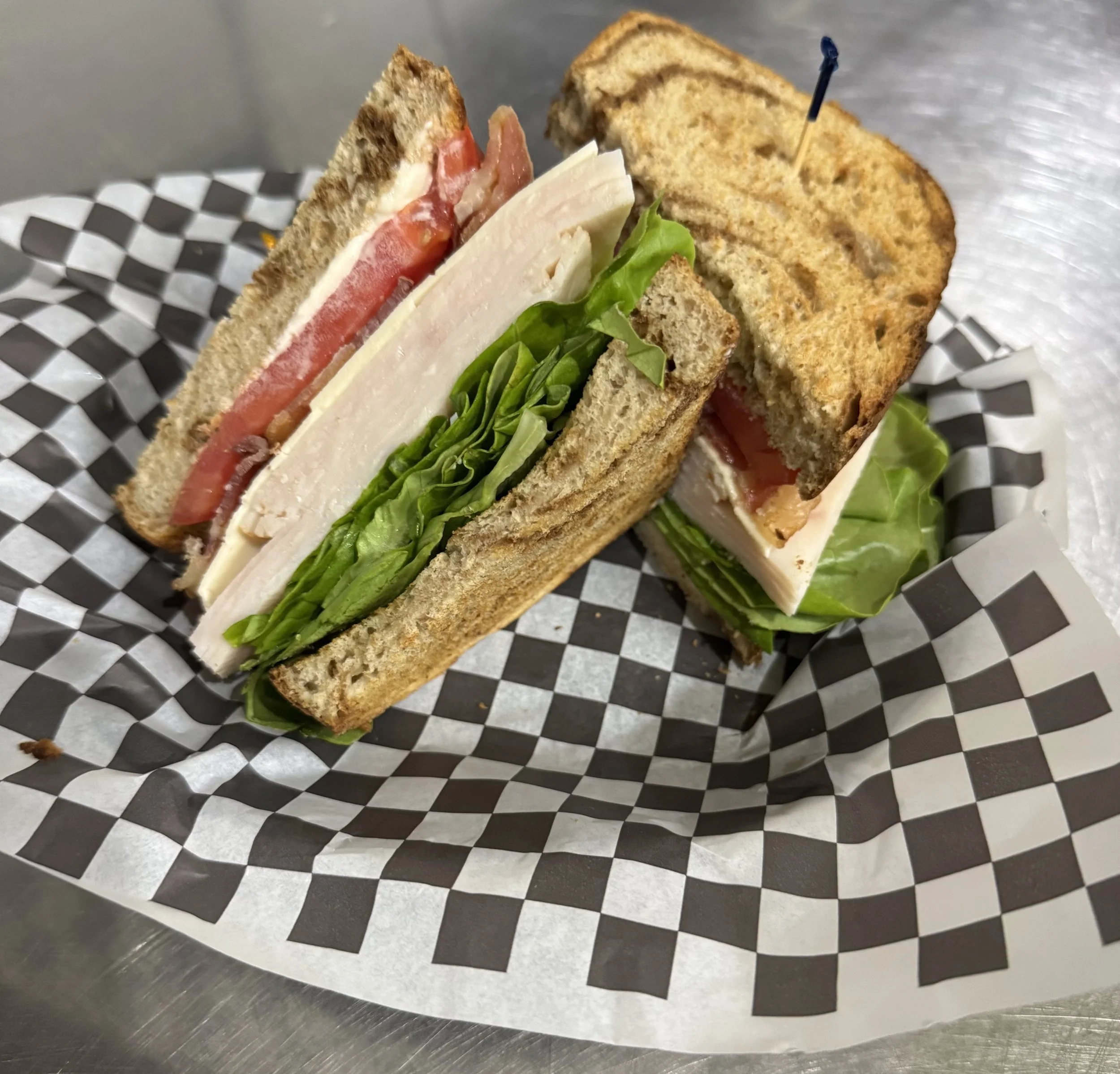 A club sandwich with layers of turkey, bacon, tomato, lettuce, and cheese on toasted bread held together with toothpicks, placed on black and white checkered paper on a metal surface.