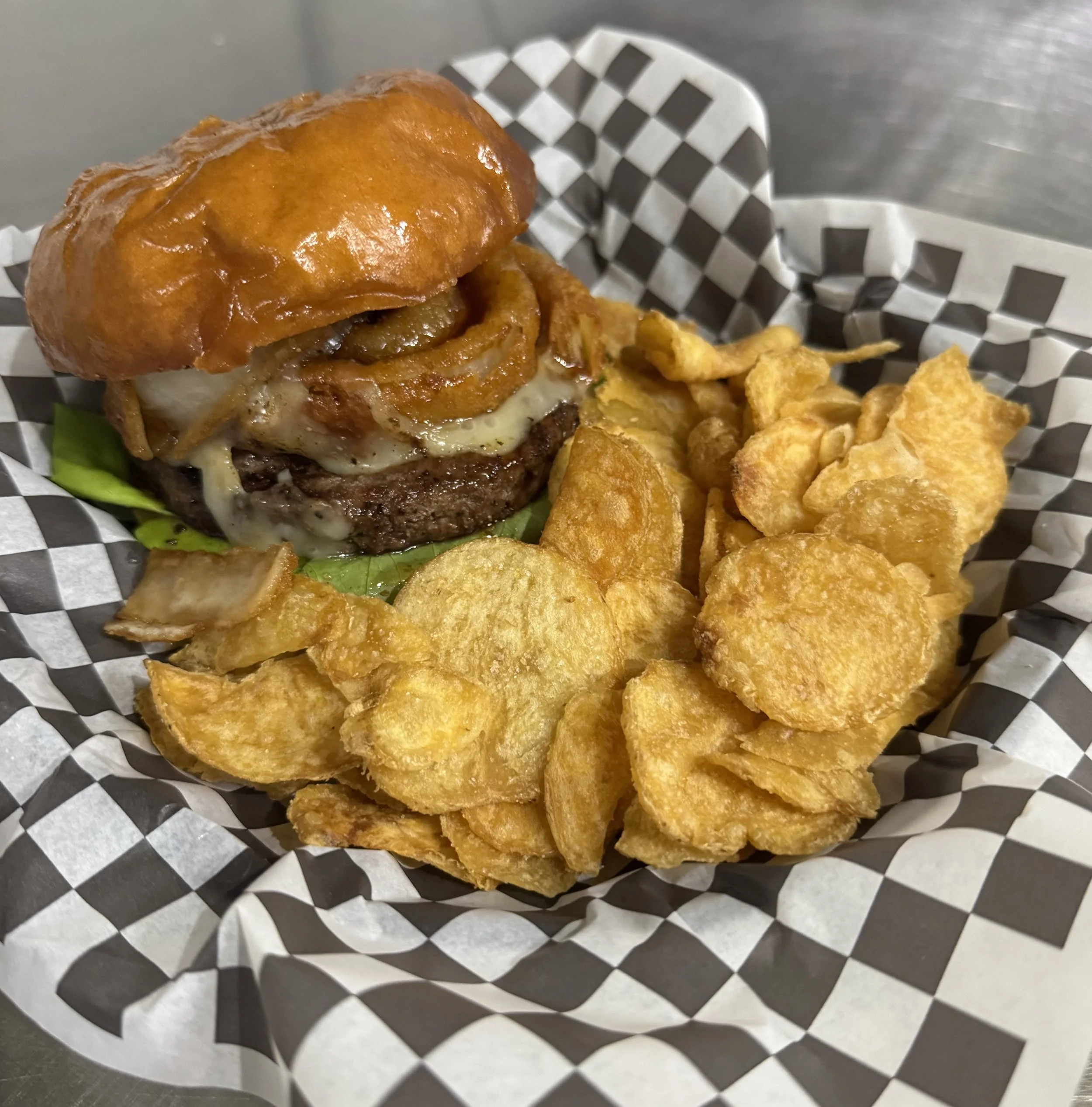 Close-up of a whiskey jack burger with lettuce, onion rings, bourbon glaze and melted pepper jack cheese, served with potato chips in checkered paper liner.