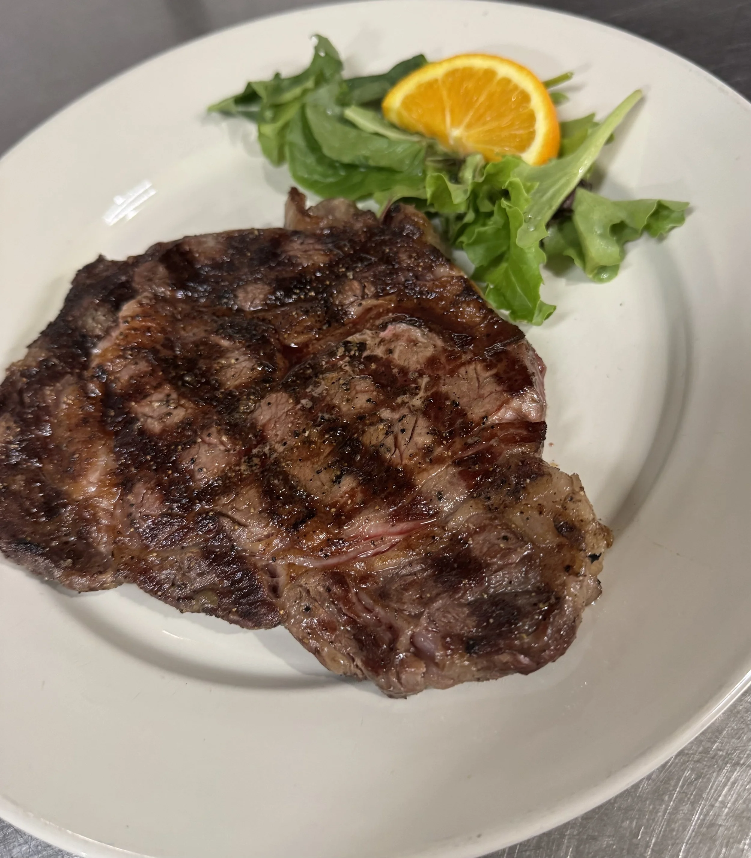 A grilled ribeye steak with grill marks served on a white plate, accompanied by a small side salad with green lettuce and a slice of orange.