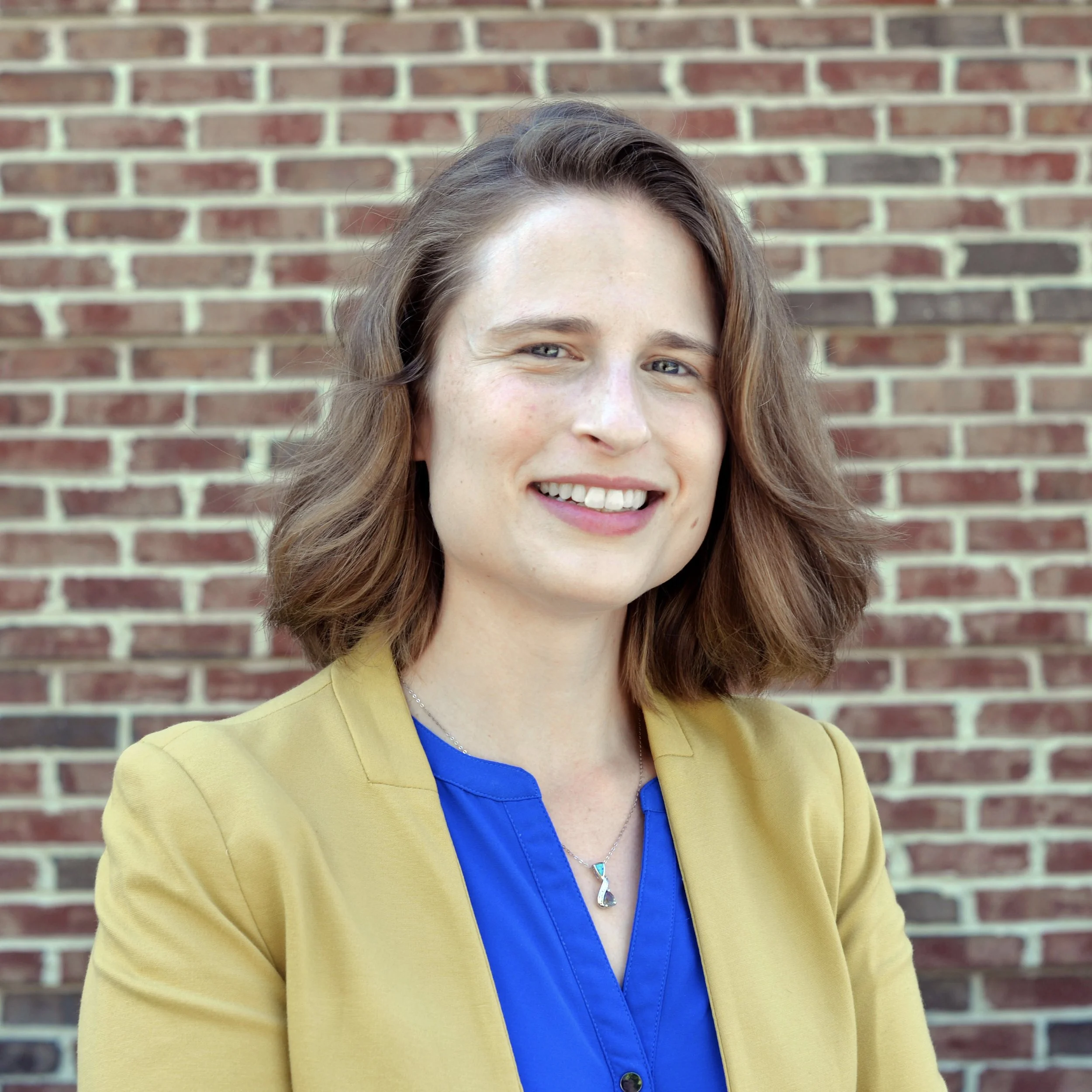 A woman with wavy, shoulder-length brown hair and light skin smiling in front of a brick wall. She is wearing a yellow blazer over a blue top with a silver necklace.