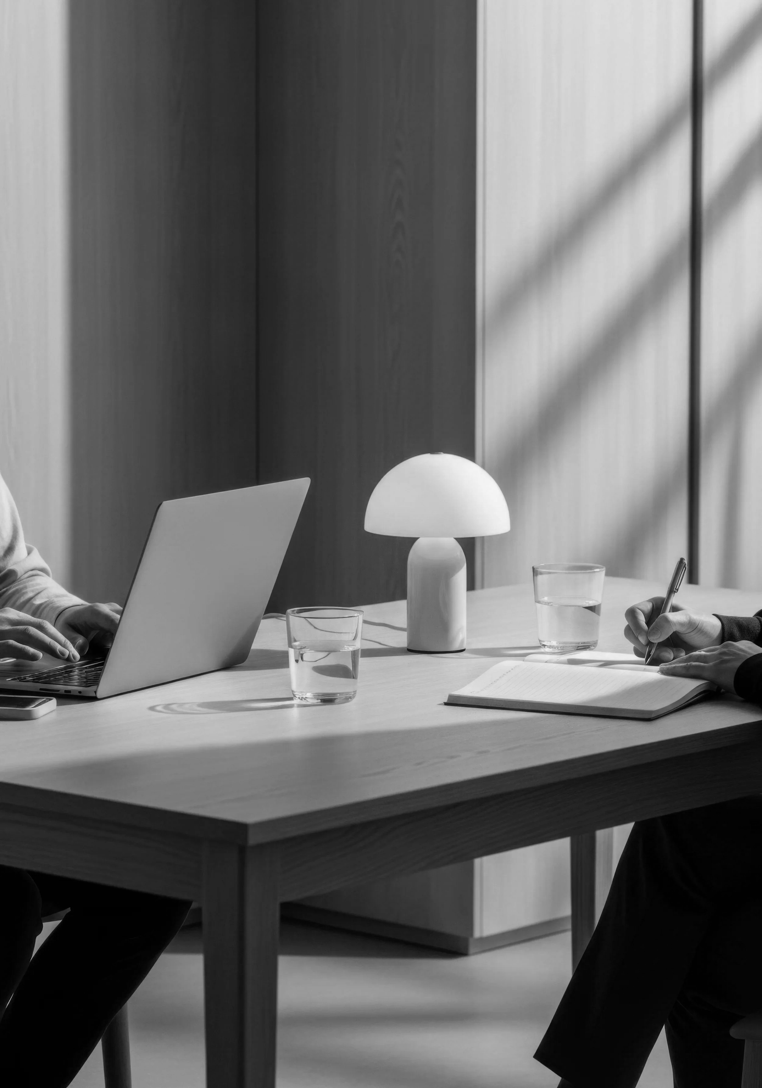 People sitting at a table working on laptops and writing in notebooks, with glasses of water and a modern desk lamp, in a professional office setting.