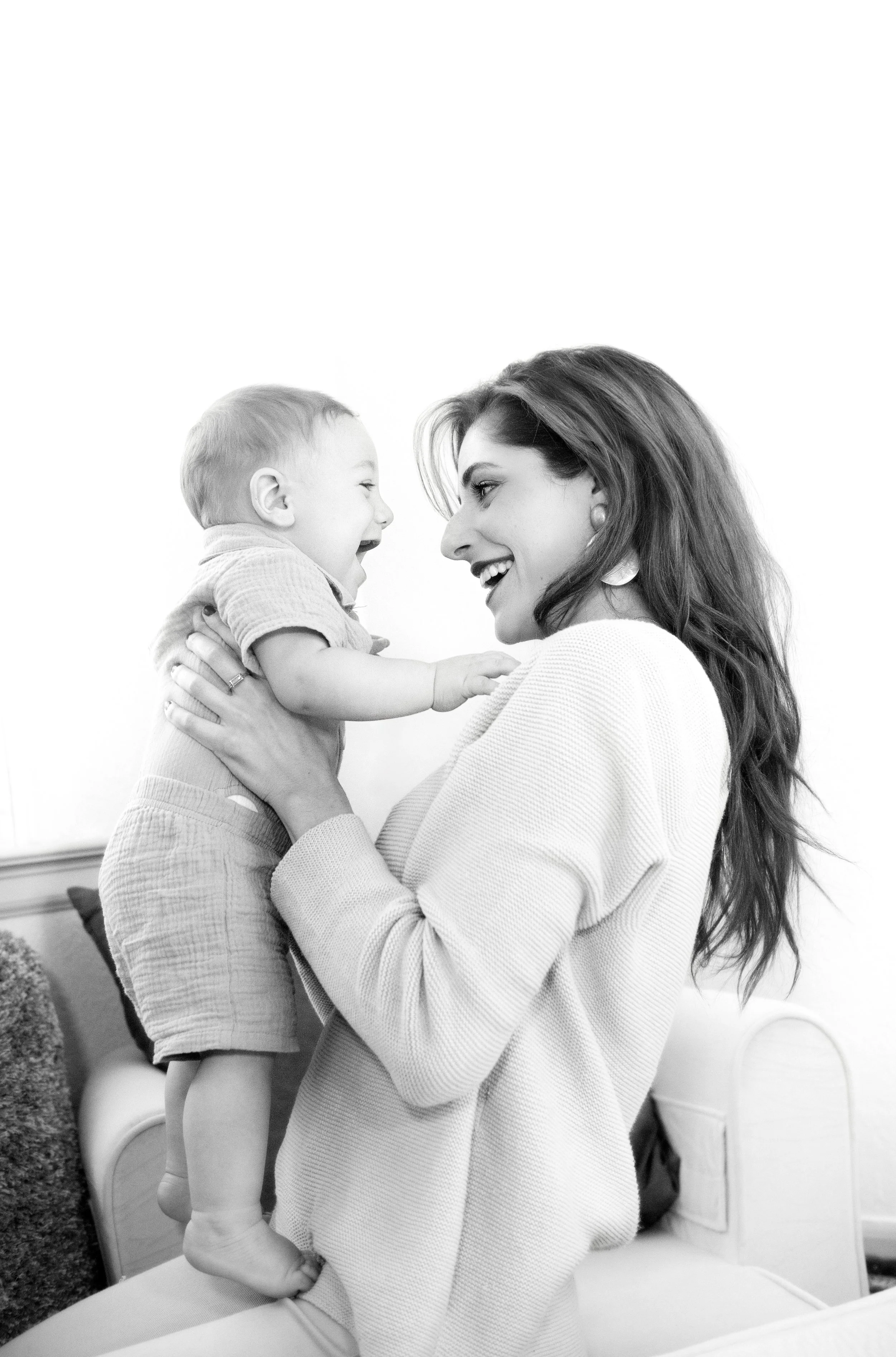 A woman holding a young boy, both smiling and facing each other, indoors with a white background.
