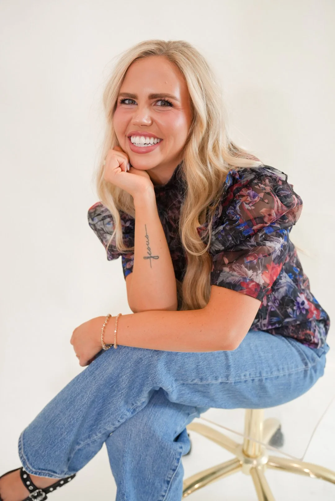 Faith is smiling with blonde hair, wearing a floral blouse and blue jeans, sitting on a chair against a plain light background.